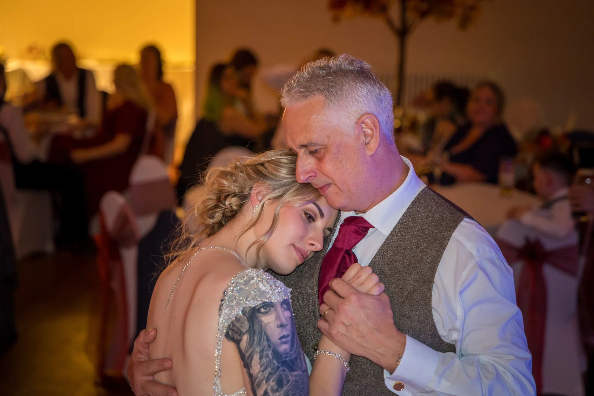 A bride with a visible arm tattoo rests her head tenderly on her father's shoulder as they share a father-daughter dance at their evening wedding reception.
