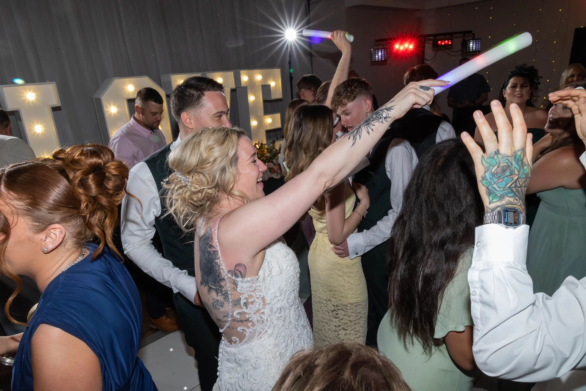 A bride with tattoos and a lace dress laughs enthusiastically on a packed dance floor illuminated by direct flash and glowing party sticks.