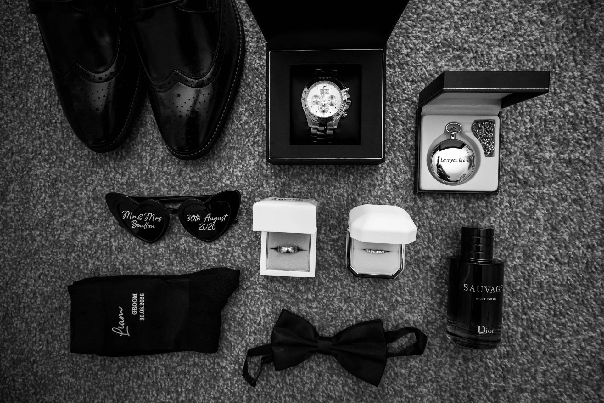 An overhead monochrome flat lay displays groom accessories including polished leather brogues, a watch, rings in boxes, a pocket watch, a fragrance bottle, sunglasses, socks, and a black bow tie.