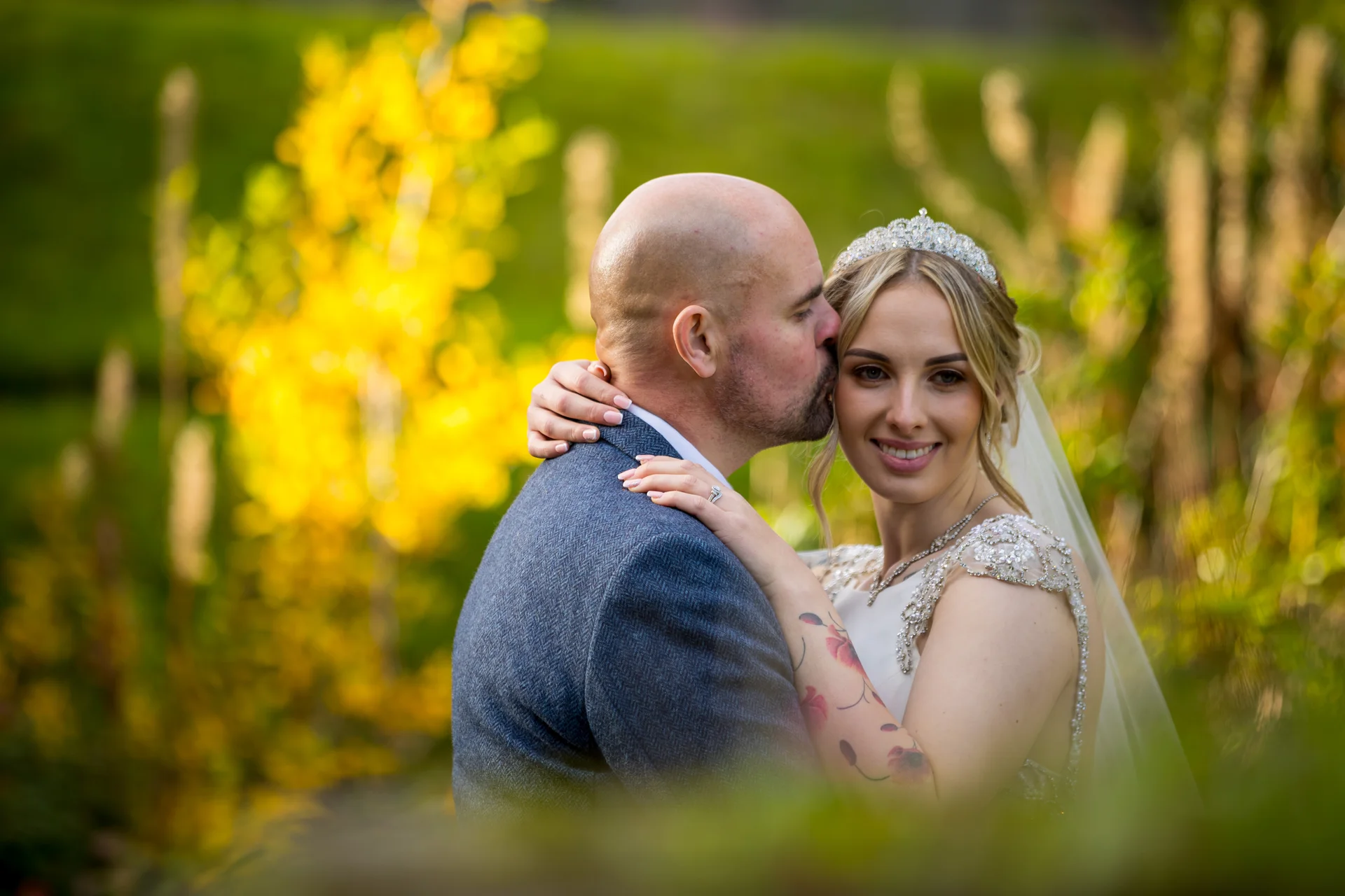 A groom gently kisses his smiling bride on the forehead outdoors amidst glowing autumn foliage during a portrait session.