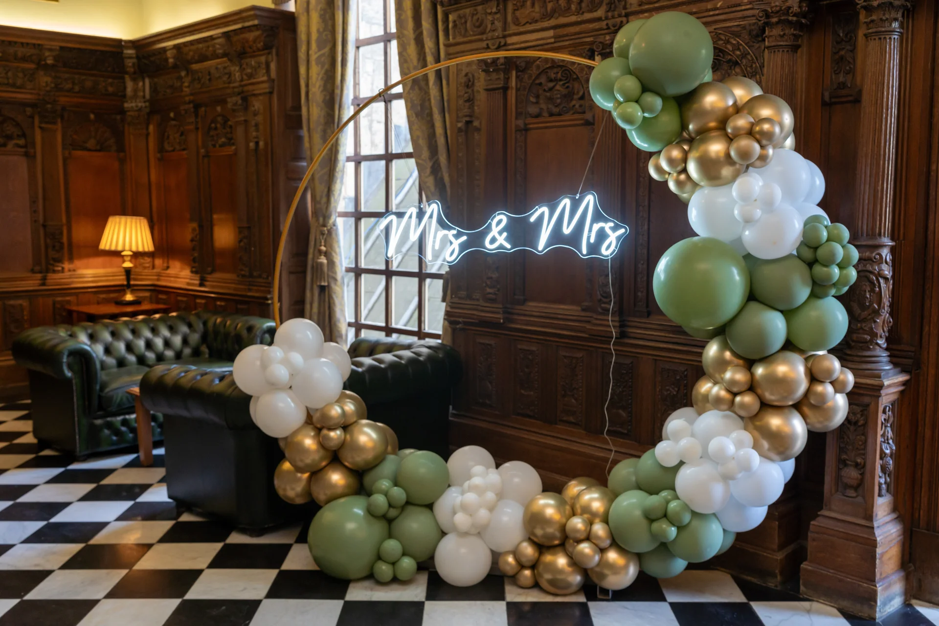 A stunning gold circular balloon hoop adorned with sage green, white, and metallic gold balloons frames an illuminated neon sign reading Mrs & Mrs inside a historic wood-panelled room.