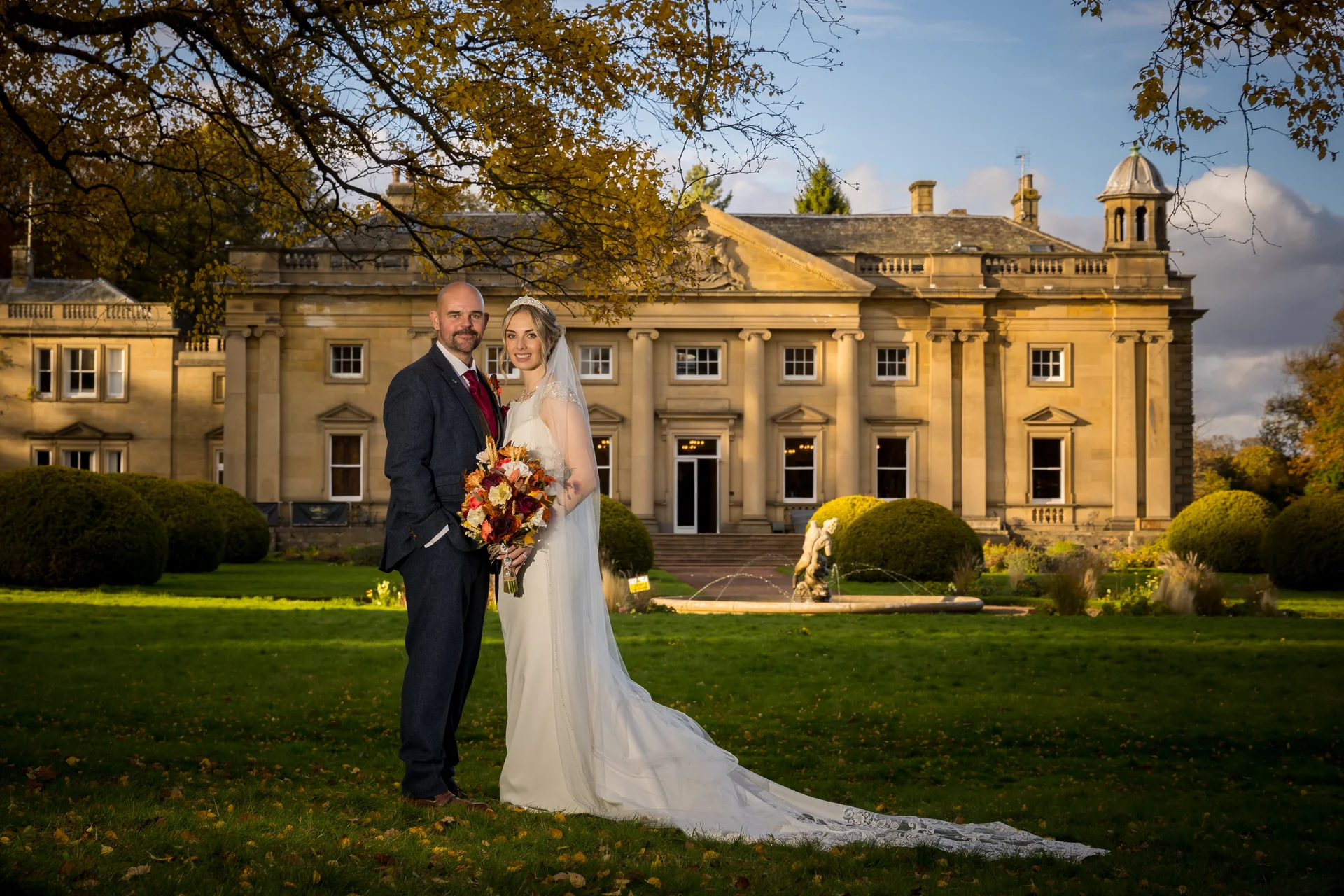 A newlywed couple poses for a portrait on the lawn in front of a grand stately home during an autumn wedding, illuminated by golden hour sunlight.