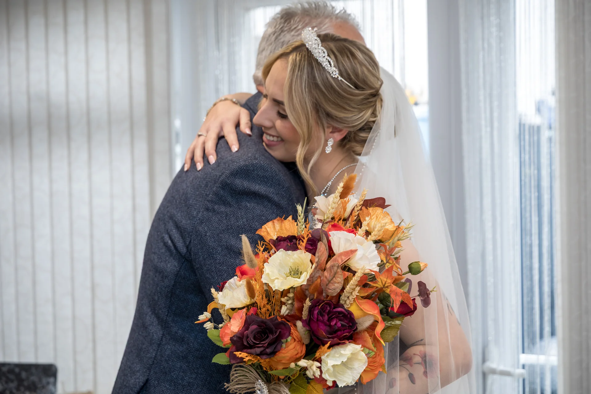 A smiling bride wearing a crystal tiara and holding a vibrant autumn-toned bouquet embraces her father in a warm, tender moment indoors.