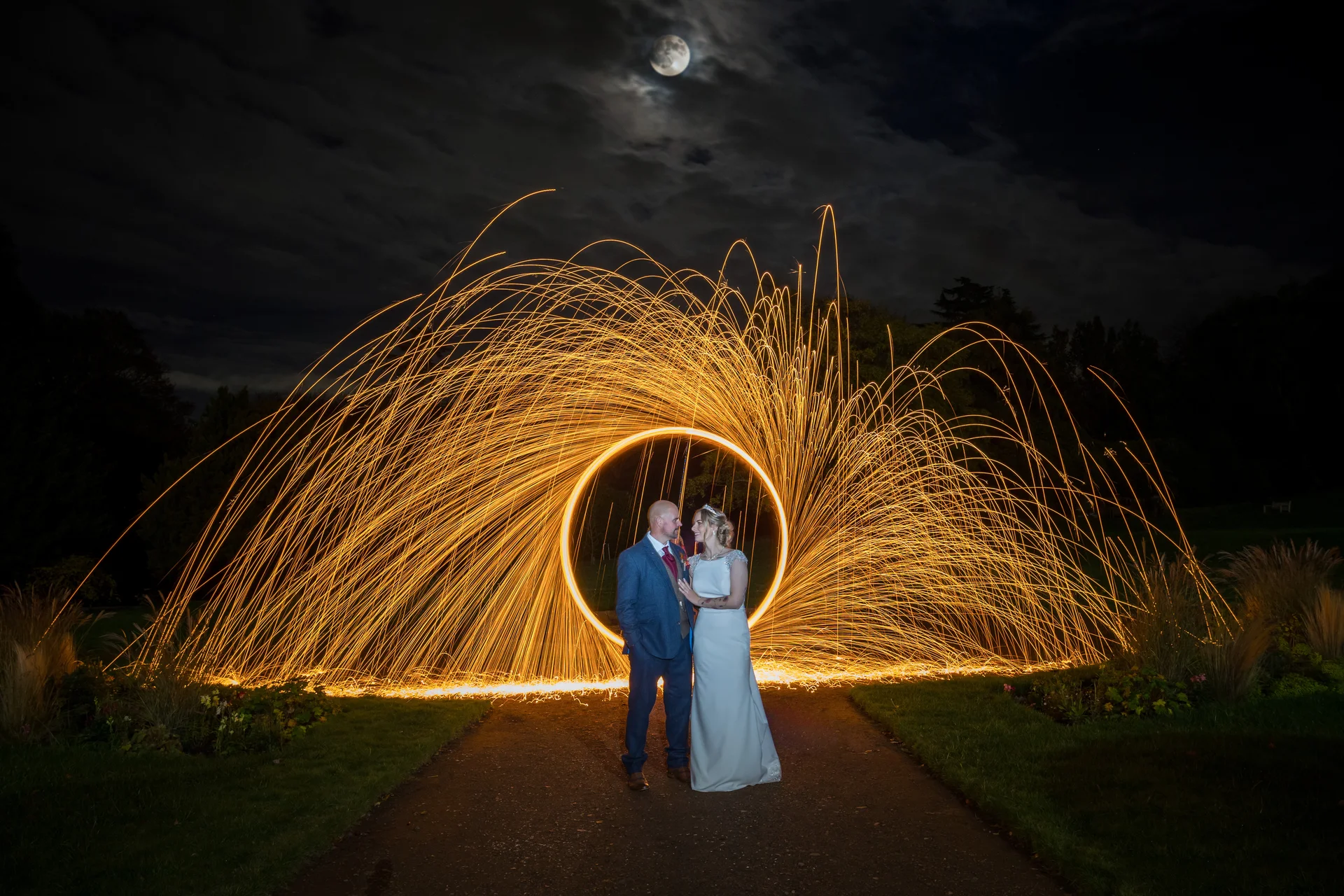 A newlywed couple stands on a pathway at night, framed by a dramatic sparkler light painting forming a glowing circle beneath a full moon.