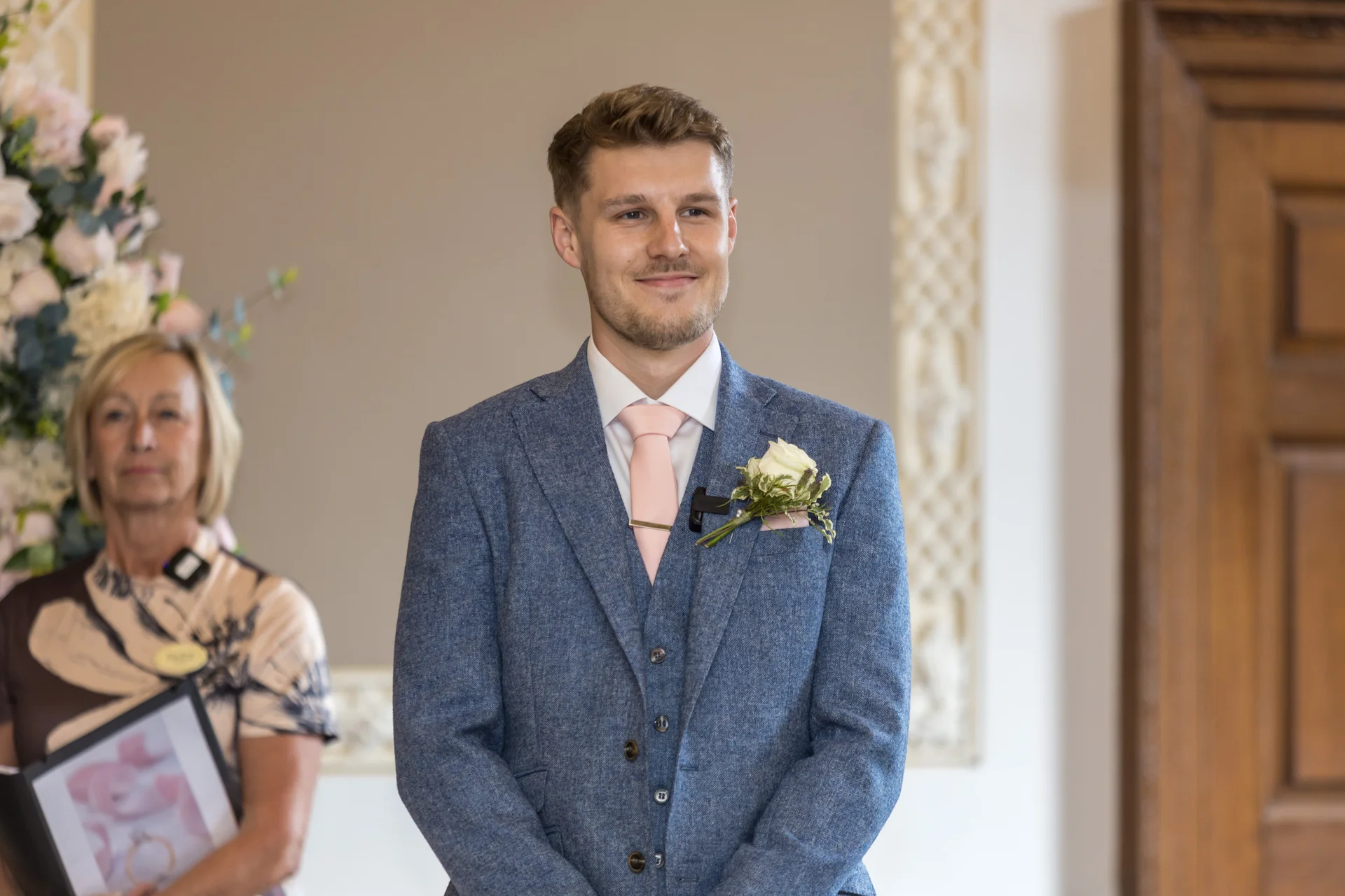 A smiling groom wearing a blue tweed three piece suit and a pink tie awaits the ceremony while standing indoors next to floral arrangements.