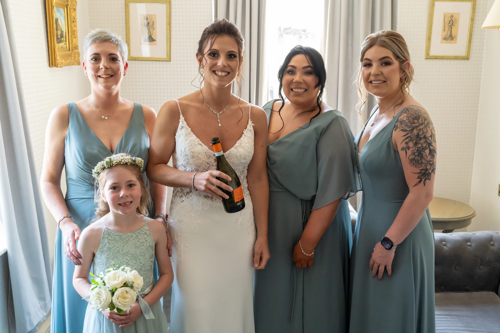 A smiling bride holding a bottle of prosecco poses indoors alongside her bridesmaids and a young flower girl wearing sage green dresses.