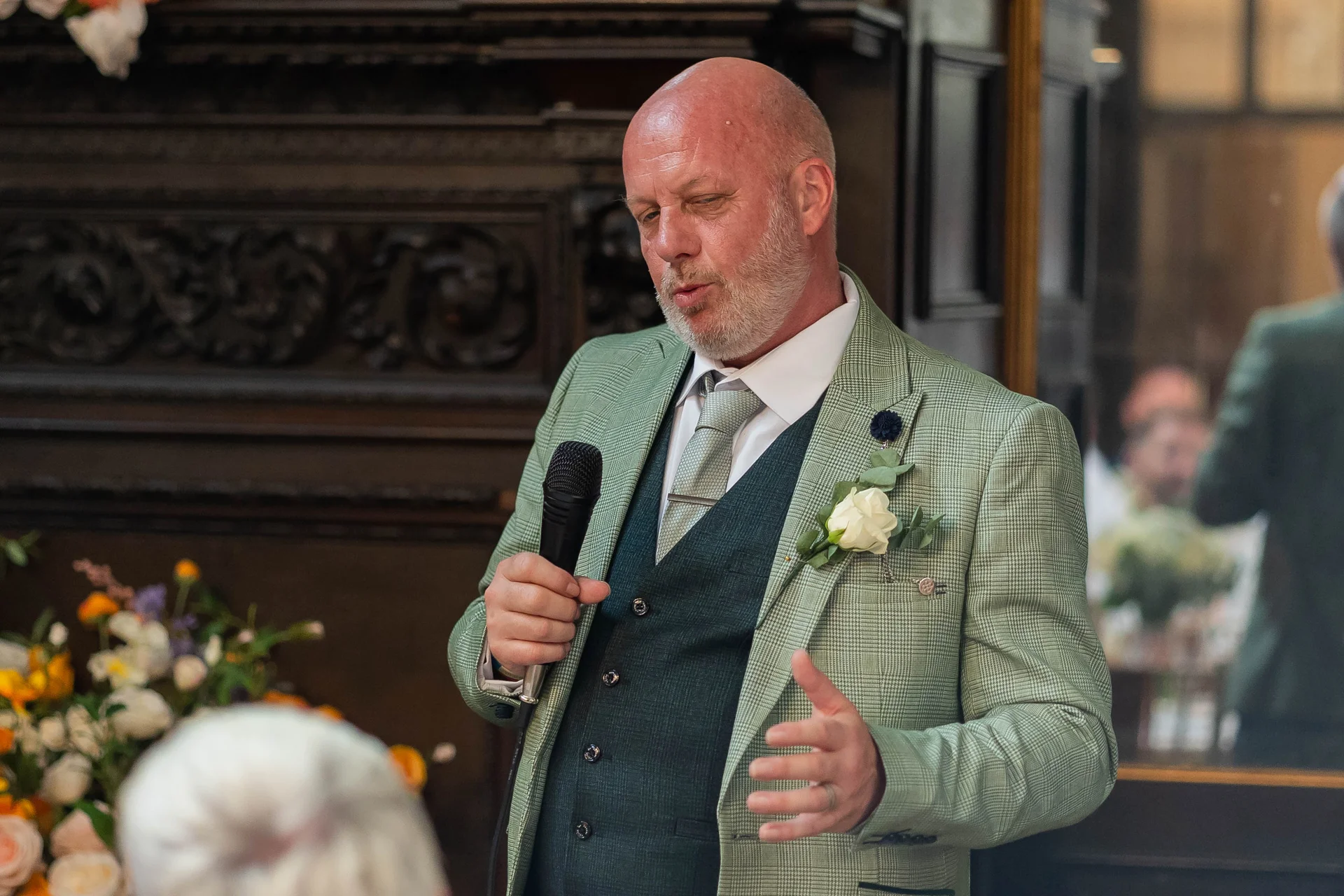 A bald groom wearing a green tweed suit and dark waistcoat delivers a speech while holding a microphone during the wedding reception.