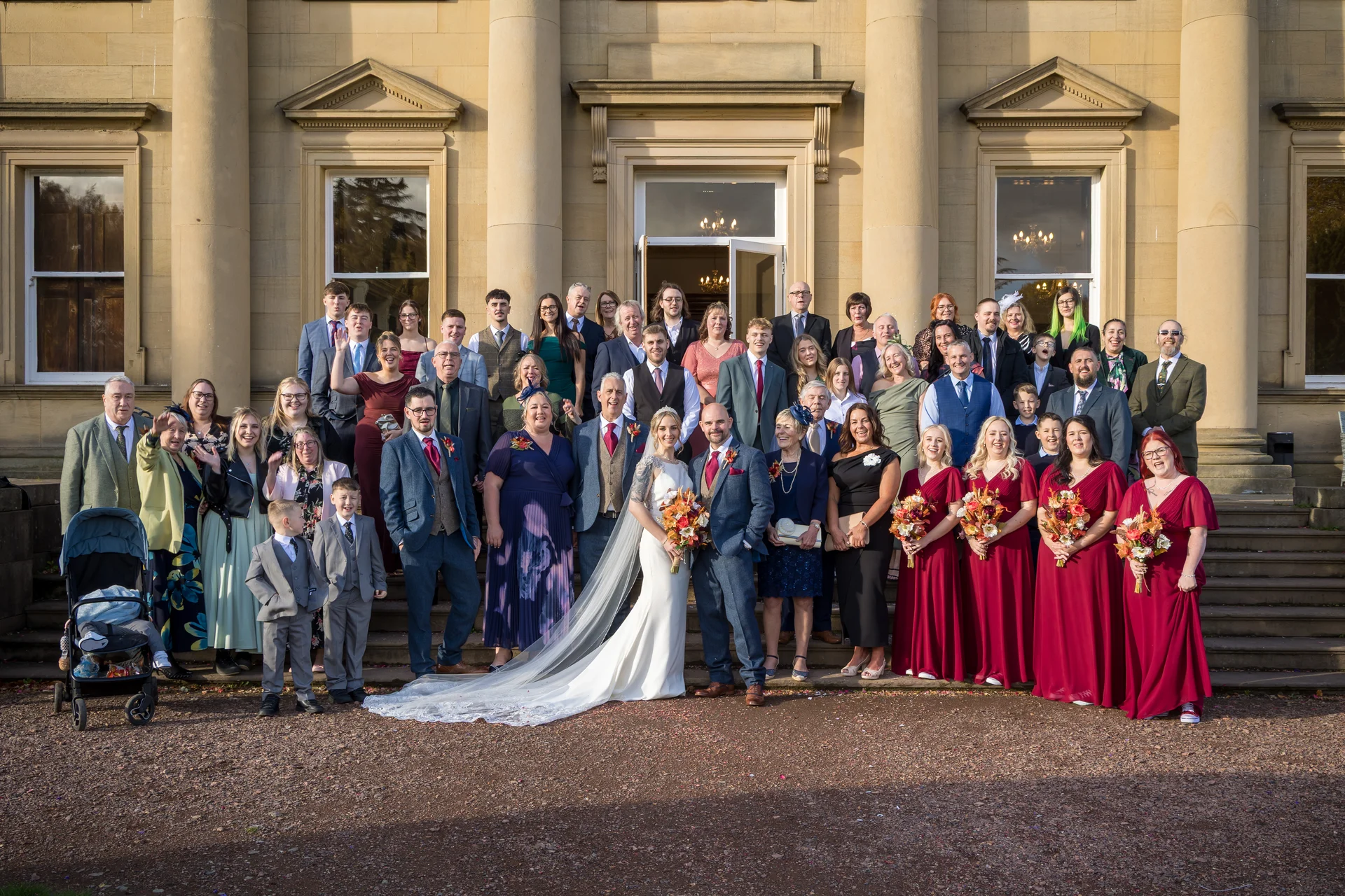 A large wedding party and family group pose on the grand stone steps outside a majestic country house, with the bride and groom at the center.