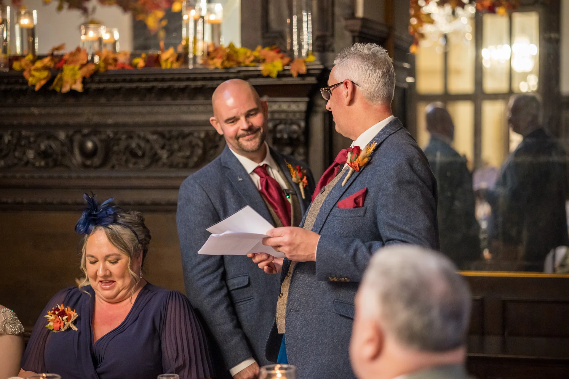 A groom in a blue tweed suit delivers his wedding speech near a decorated fireplace while guests look on and laugh during the reception.