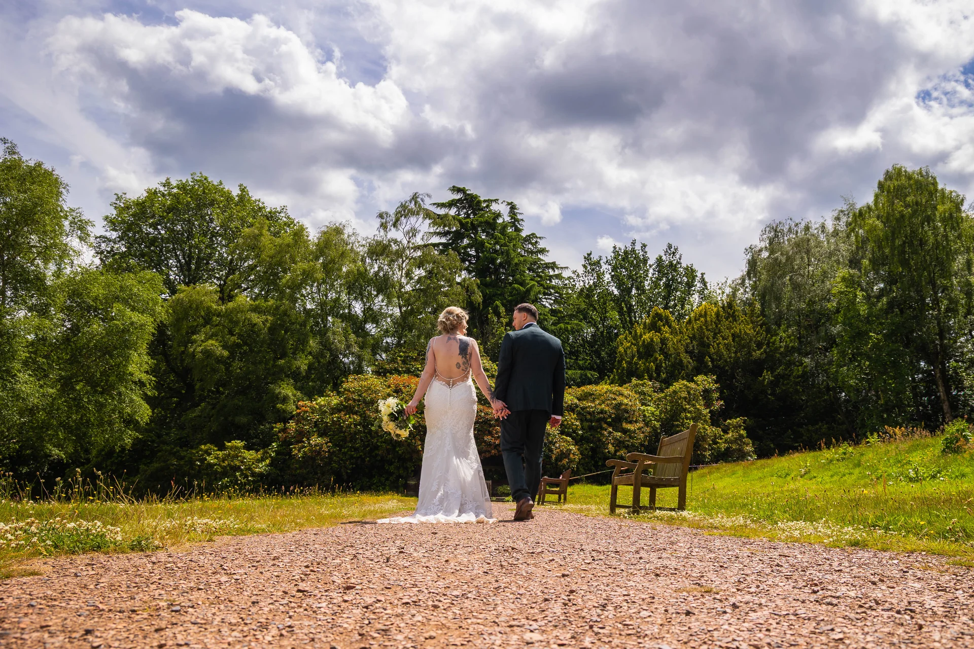 Newlyweds walk hand in hand down a gravel path away from the camera, framed by lush green trees and a dramatic cloudy sky.