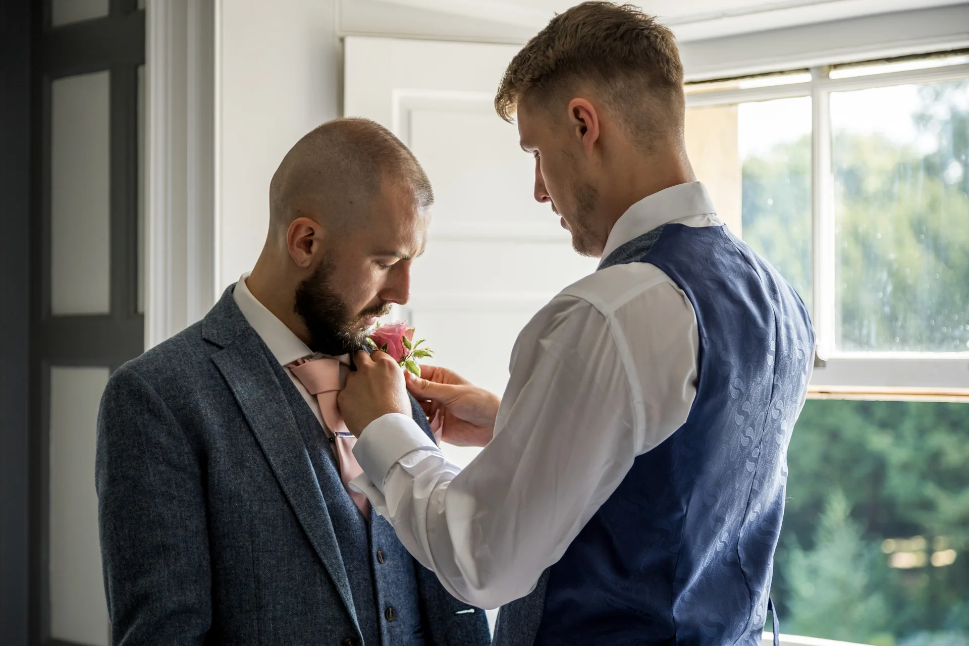 One groom gently pins a floral boutonniere onto his partner's lapel in a brightly lit room during morning preparations.