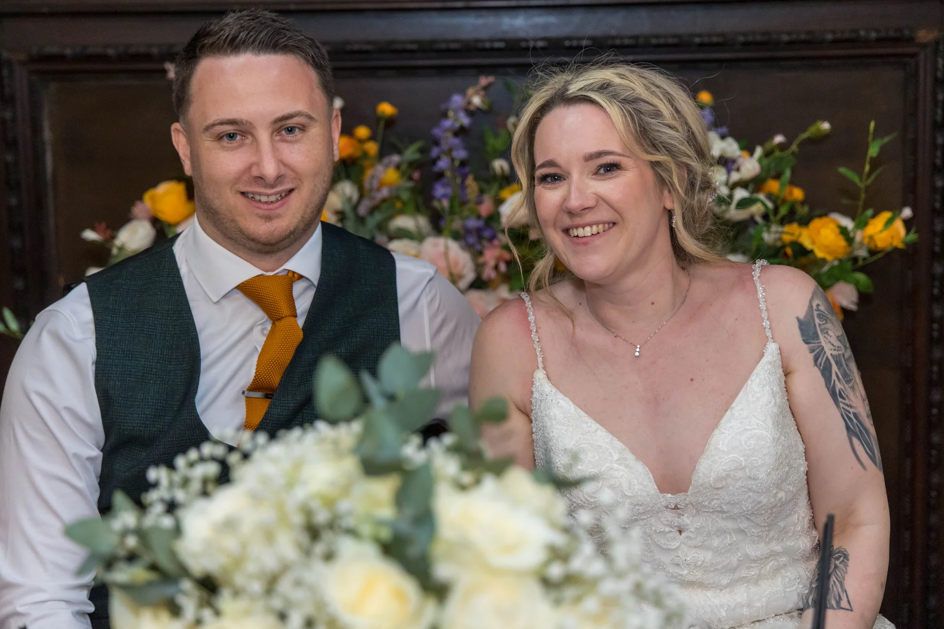 A smiling bride and groom sit together at the top table, framed by floral centrepieces with white roses and vibrant yellow blooms.