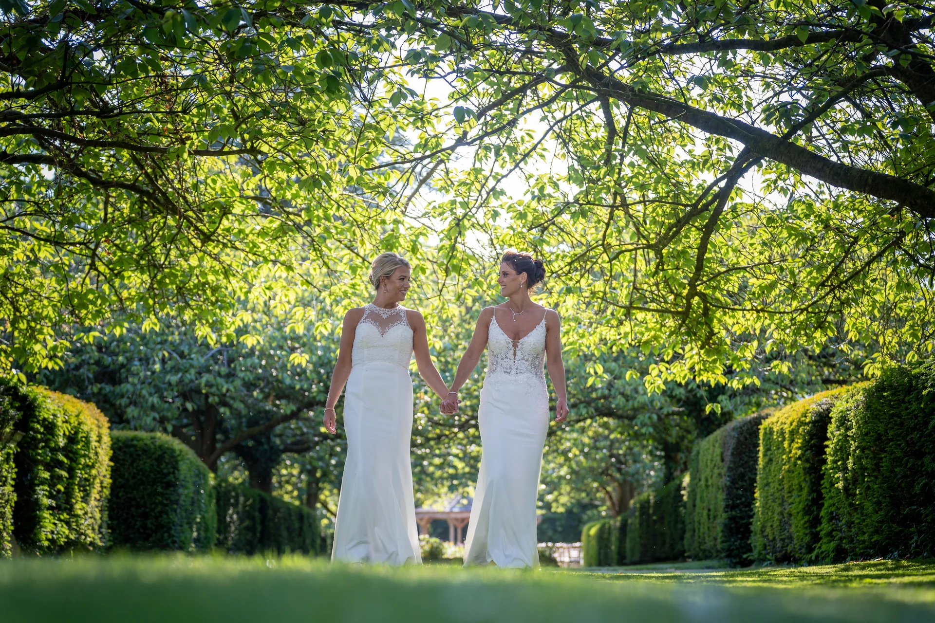 Two brides in elegant white wedding dresses walk hand in hand through a sunlit, tree-lined garden path, looking lovingly at each other.