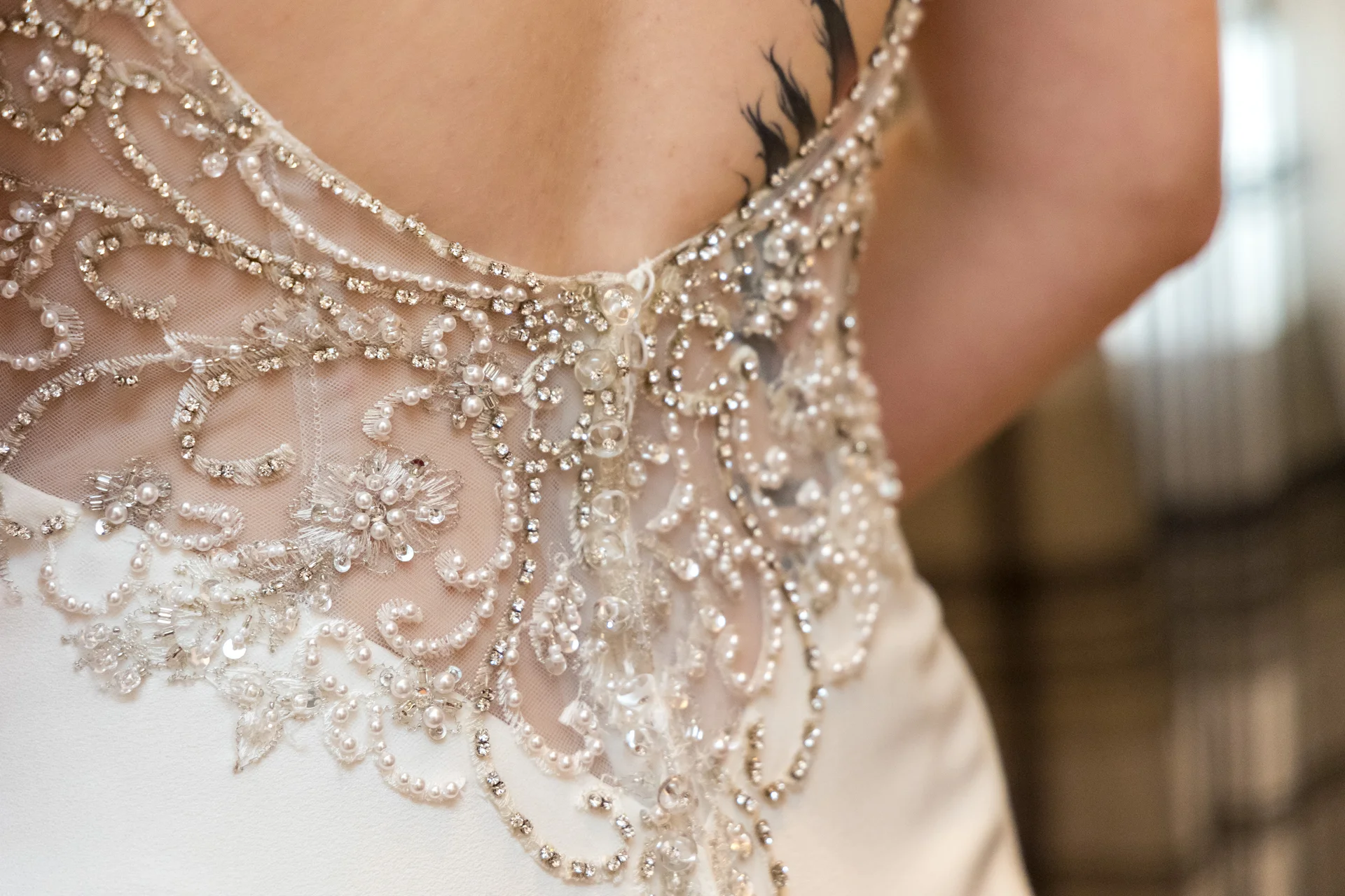 Close-up of the intricately beaded and pearl-embellished low open back of a bride's wedding gown revealing a visible shoulder tattoo.