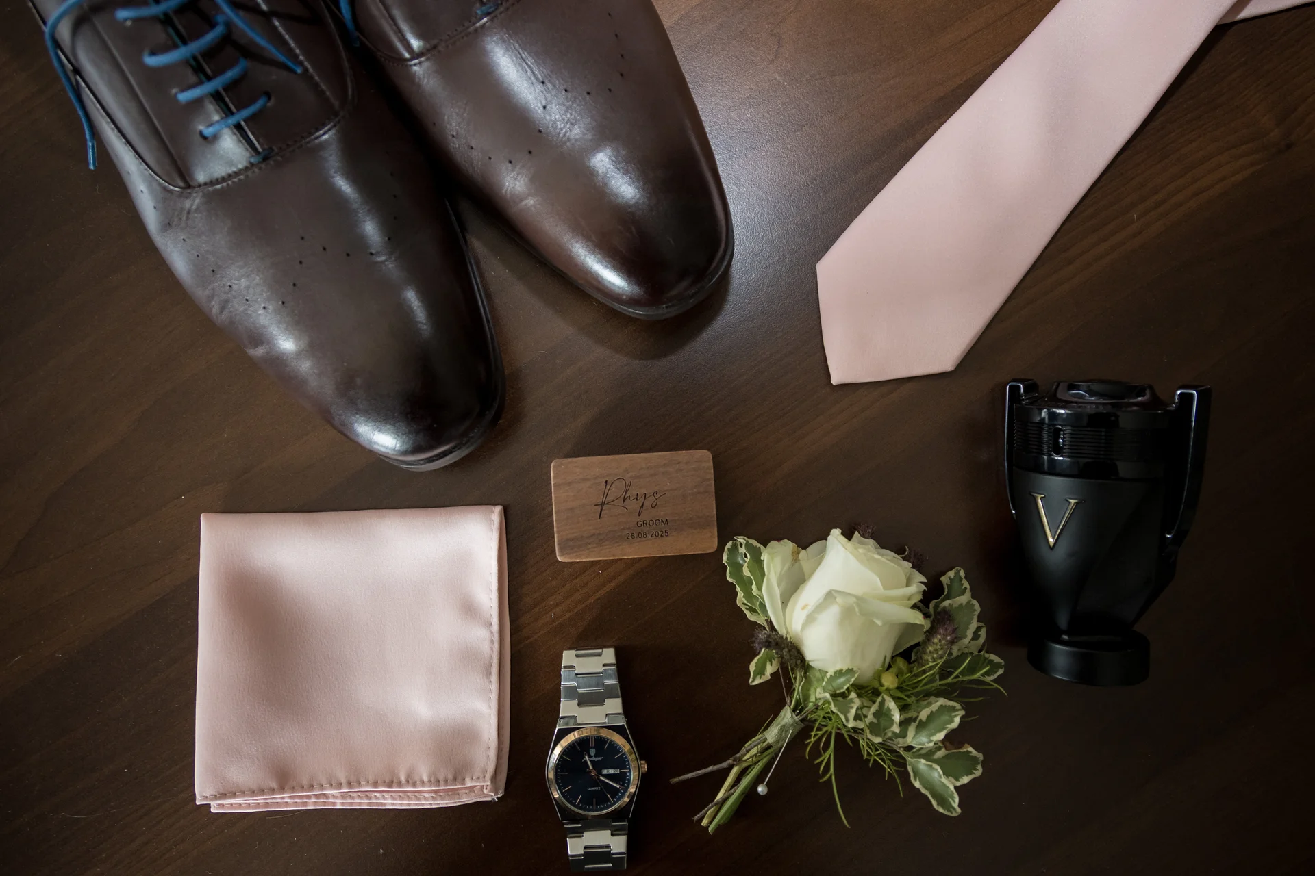 A flat lay of groom accessories on a wooden table, featuring brown leather shoes, a pink tie and pocket square, a watch, a white rose boutonniere, and a bottle of aftershave.