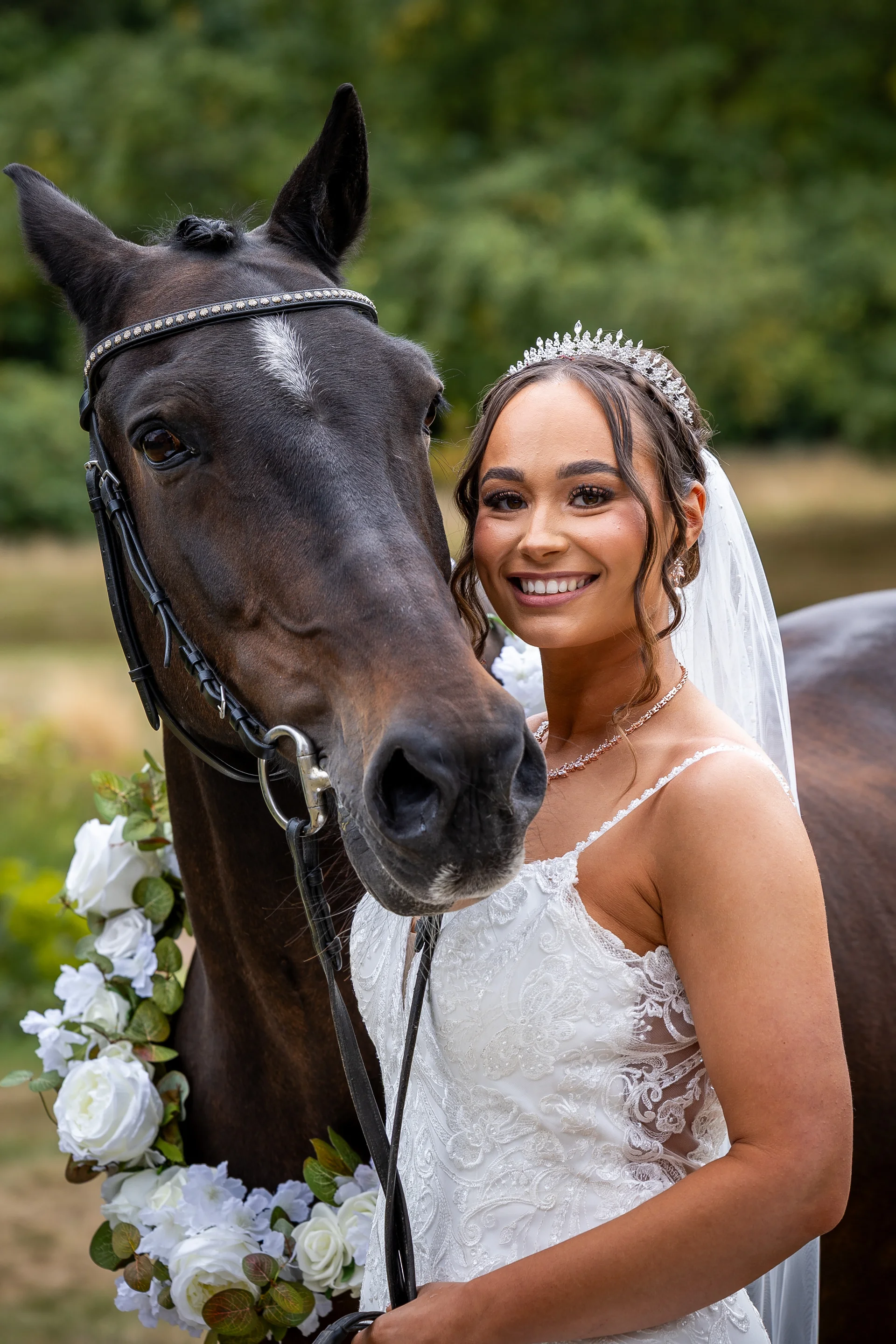 A smiling bride wearing a crystal tiara and lace wedding dress poses outdoors next to a dark horse adorned with a white floral collar.