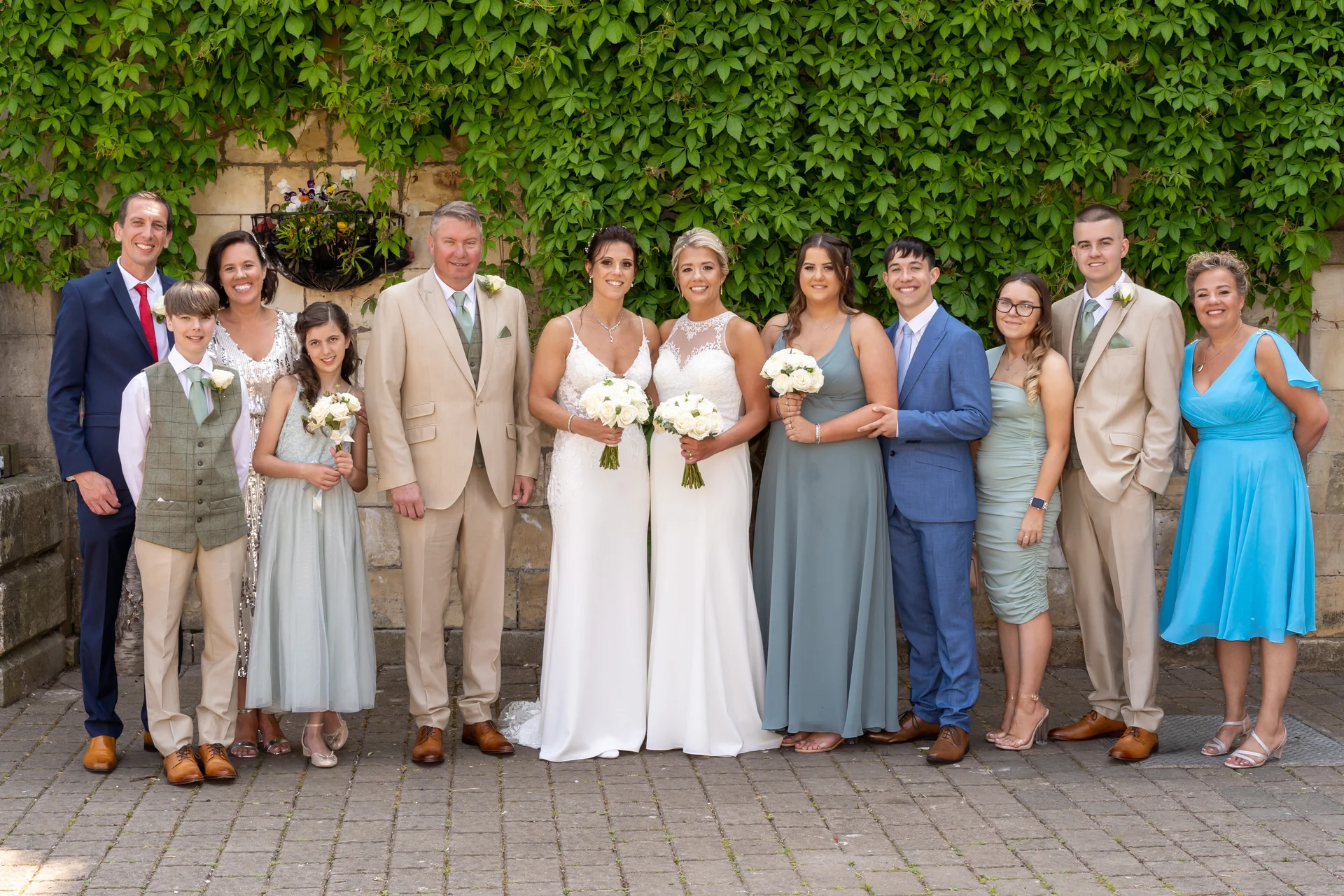 A full-length group portrait of two brides in white gowns standing alongside their wedding party, featuring groomsmen in beige and blue suits and bridesmaids in sage and dusty blue dresses against an ivy-covered stone wall.