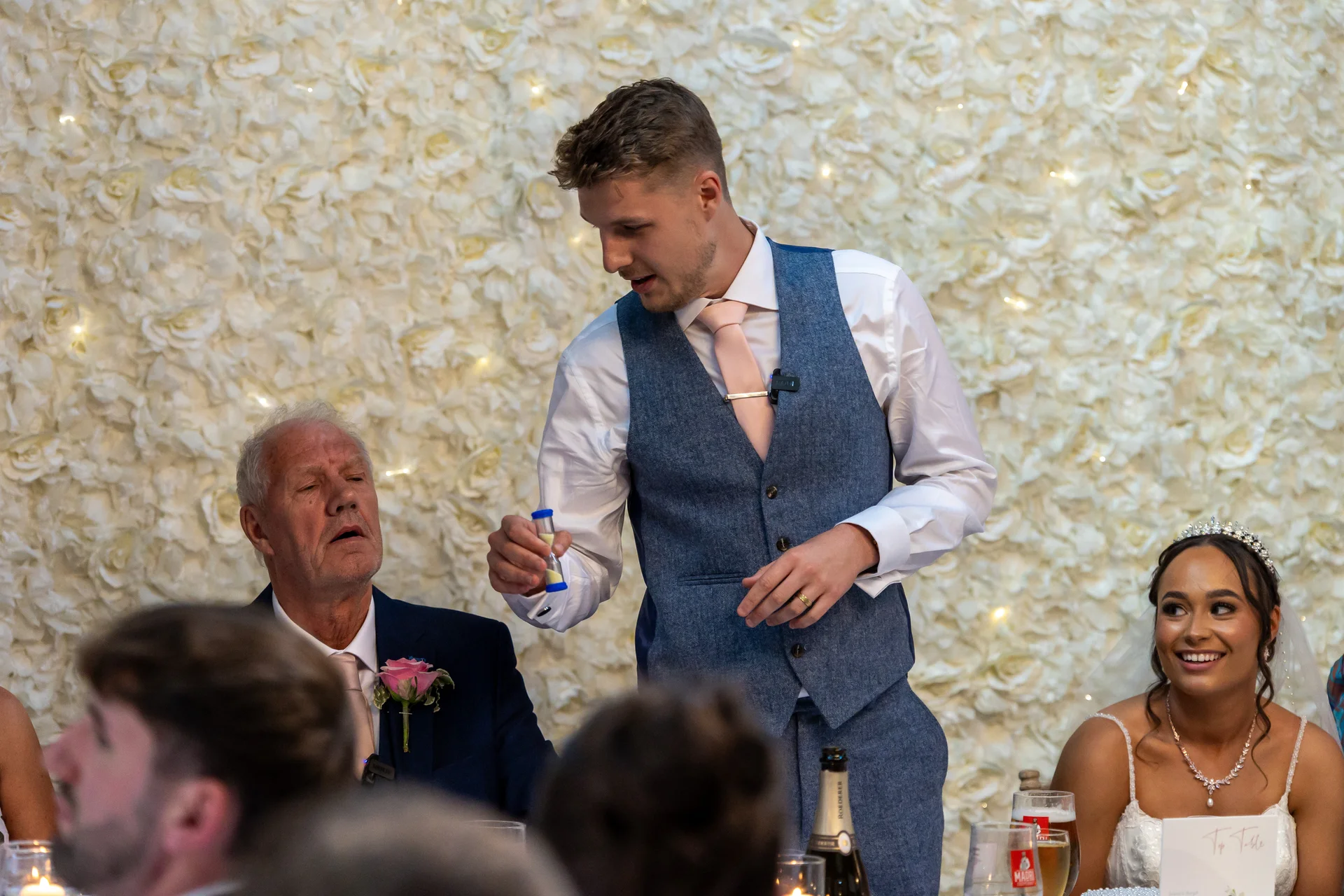 A magician in a blue waistcoat stands by a head table in front of a white flower wall, performing a trick for guests while a smiling bride looks on.