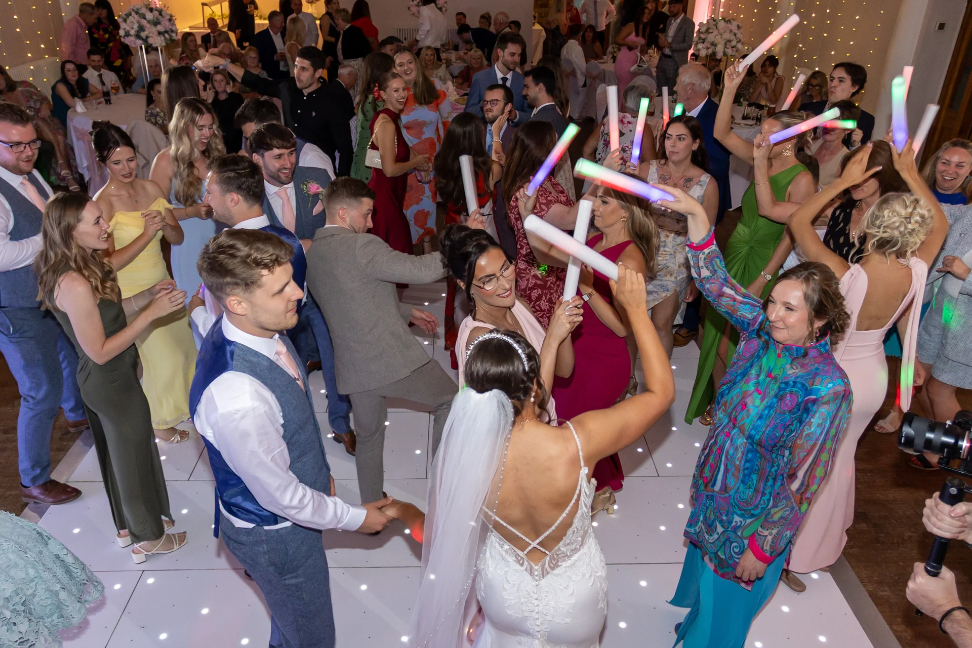 The bride and groom share a joyful moment dancing on a glowing white illuminated dance floor surrounded by wedding guests waving glowing batons.