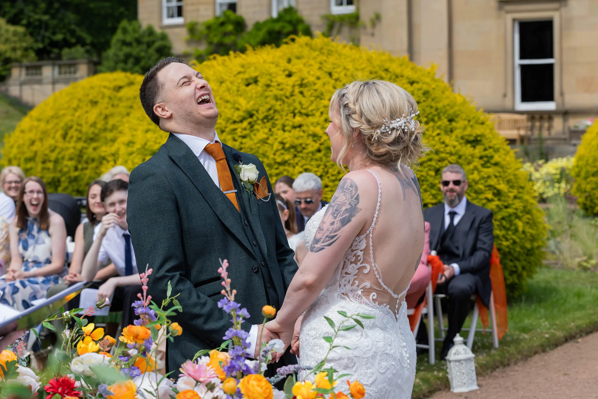 The groom throws his head back in joyful laughter while holding hands with his bride during their outdoor wedding ceremony in front of seated guests.