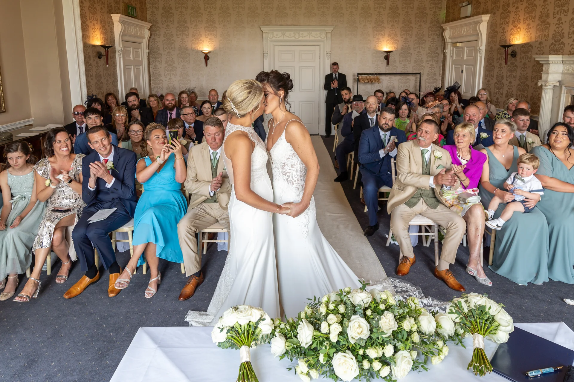 Two brides share their first kiss as a married couple at the front of a light-filled ceremony room while seated guests applaud.