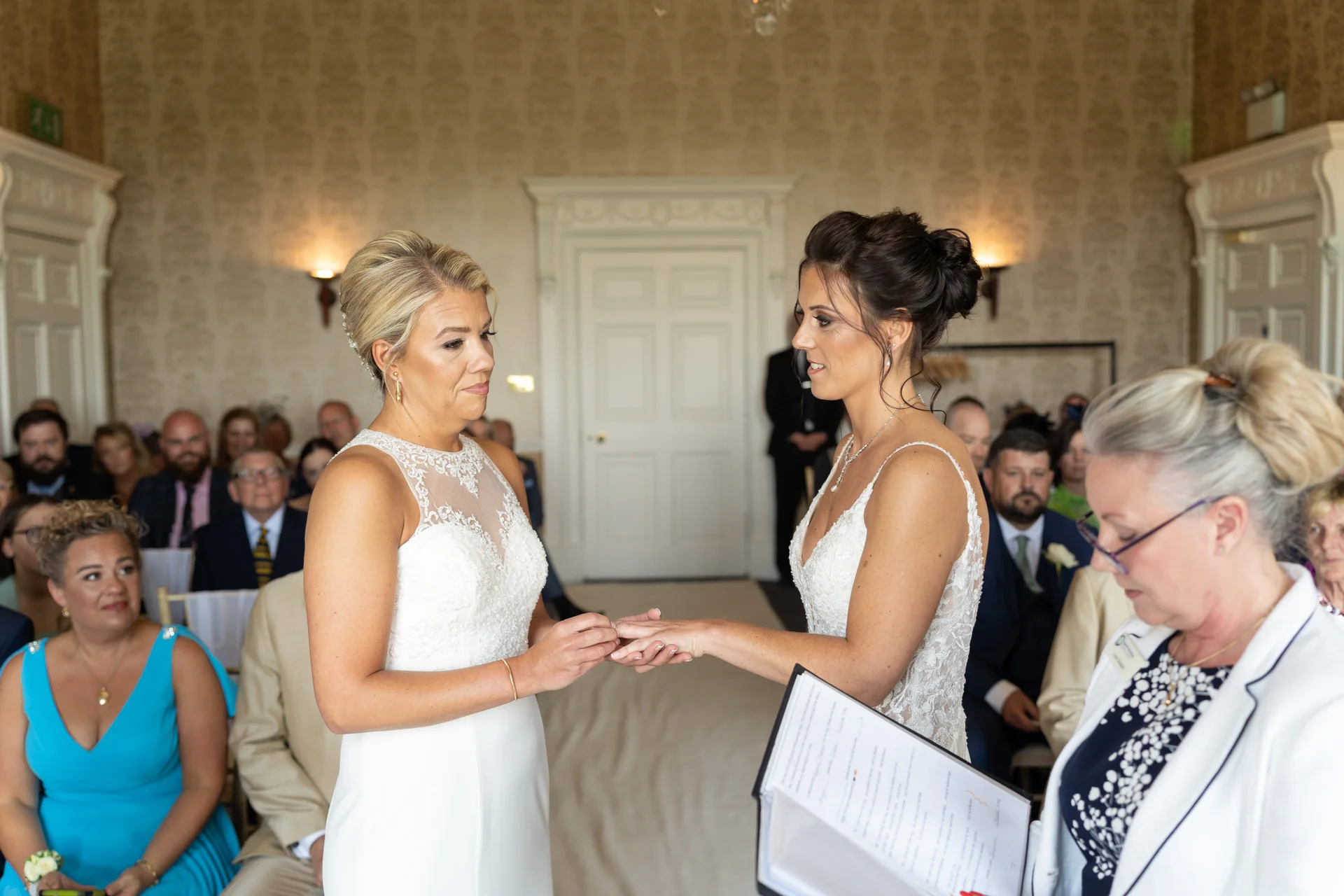 Two brides exchange rings during an indoor civil ceremony inside a light-filled manor house reception room while guests watch from seating rows.
