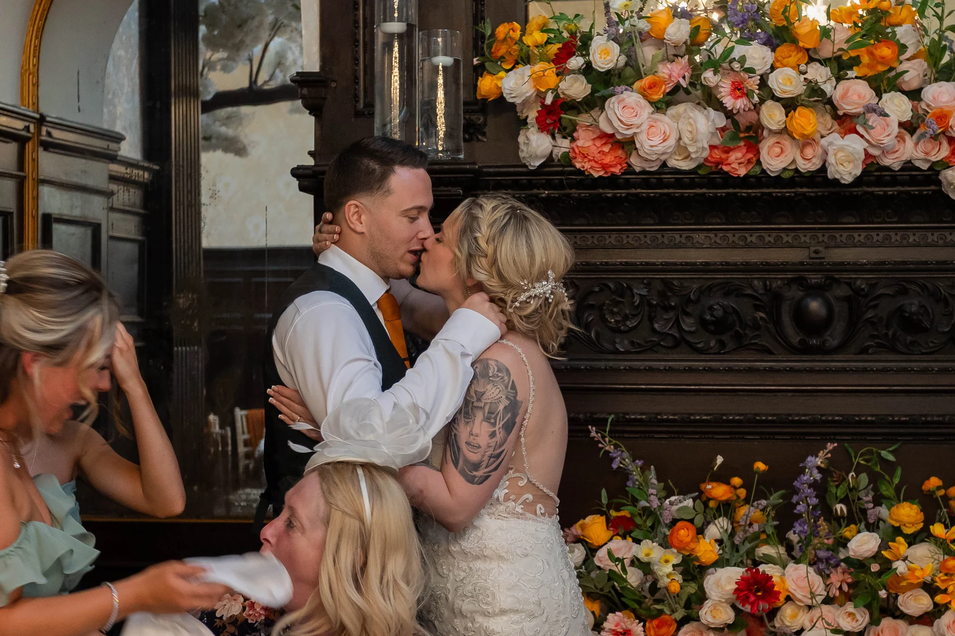 A bride with a shoulder tattoo and a groom in a waistcoat share a tender kiss in front of an ornate fireplace decorated with vibrant orange, pink, and white floral arrangements.