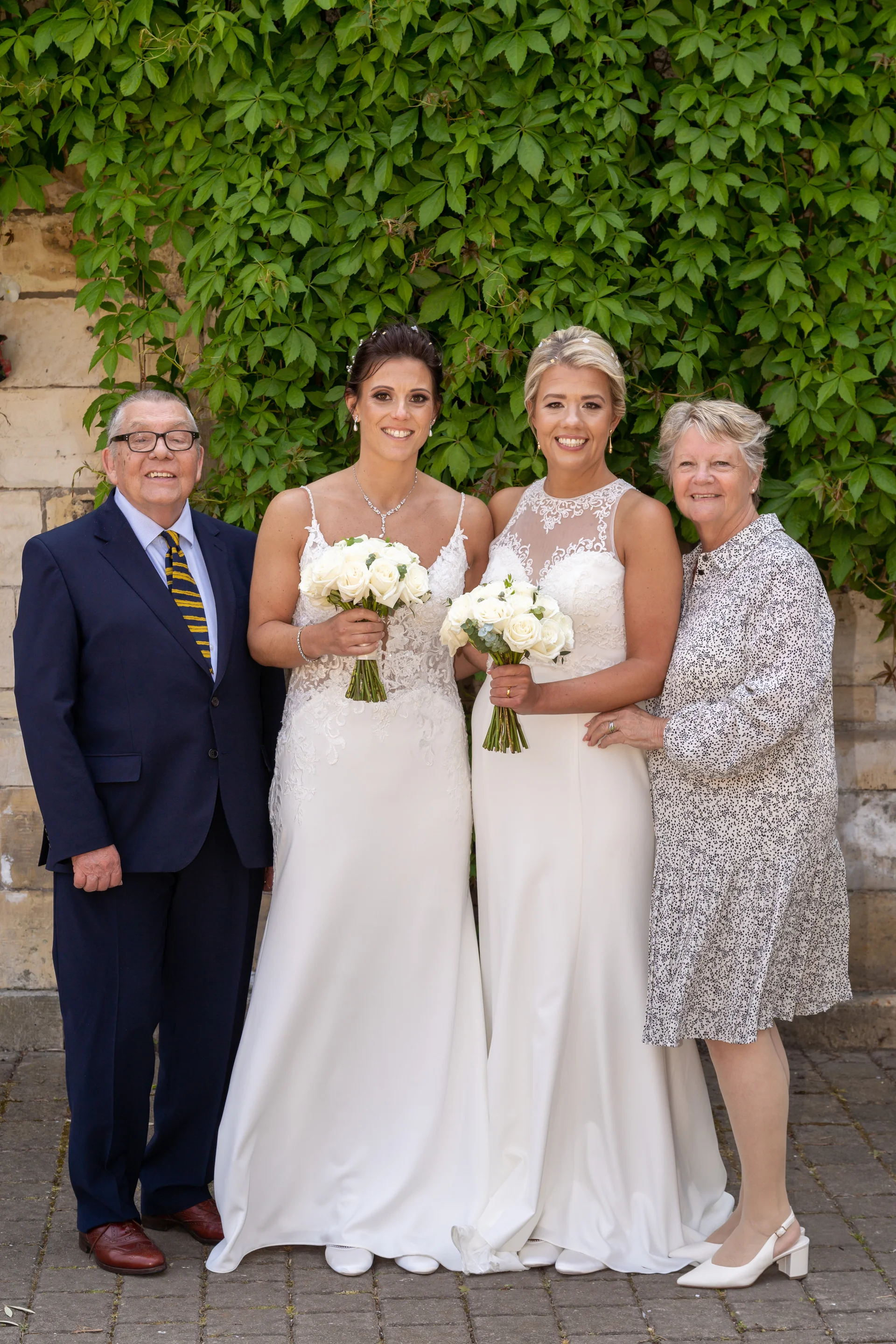 A formal family portrait of two brides in white wedding dresses holding white rose bouquets, standing alongside an older man in a navy suit and a woman in a patterned dress against an ivy-covered stone wall.