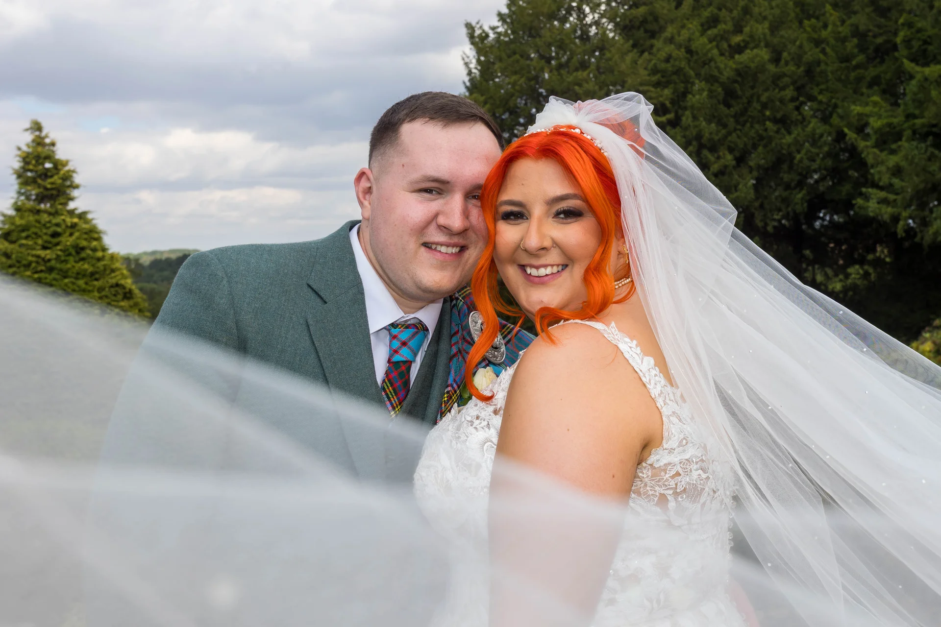 A smiling bride with vibrant orange hair and a lace wedding dress poses outdoors alongside her groom in a green suit, partially framed by a flowing white veil in the foreground.