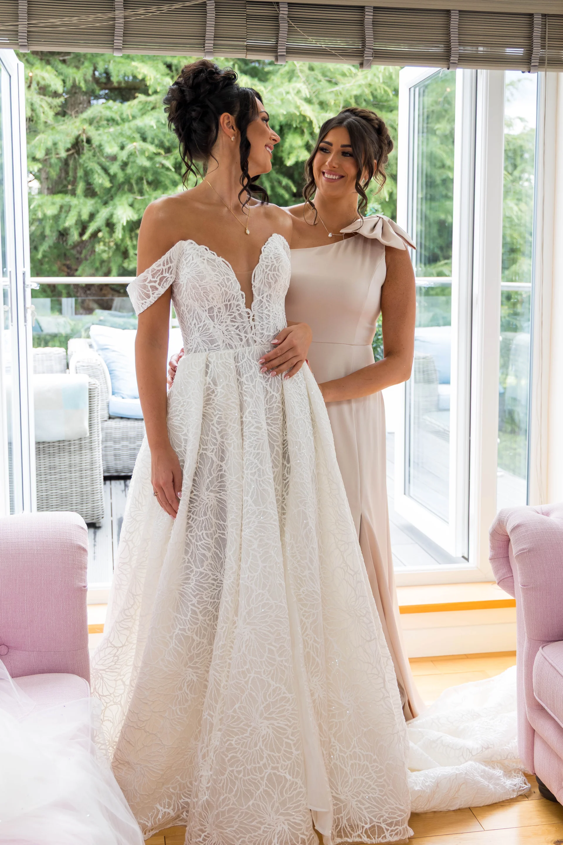 A smiling bride in an intricate lace off-the-shoulder gown stands indoors next to a bridesmaid wearing a one-shoulder champagne dress, framed by bright window light.