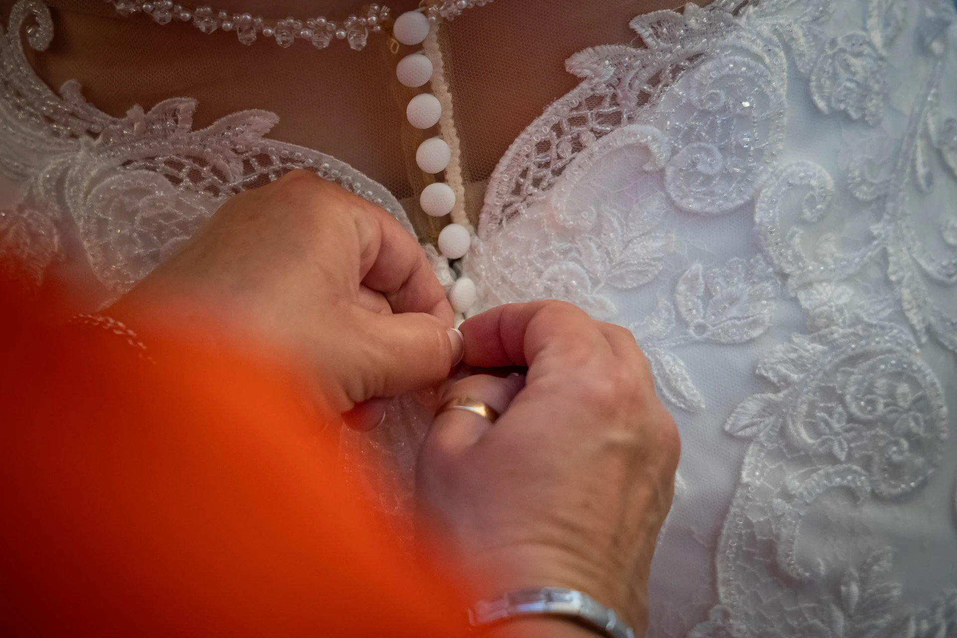 Close-up of hands fastening the intricate fabric buttons along the sheer illusion lace back of a white bridal gown.