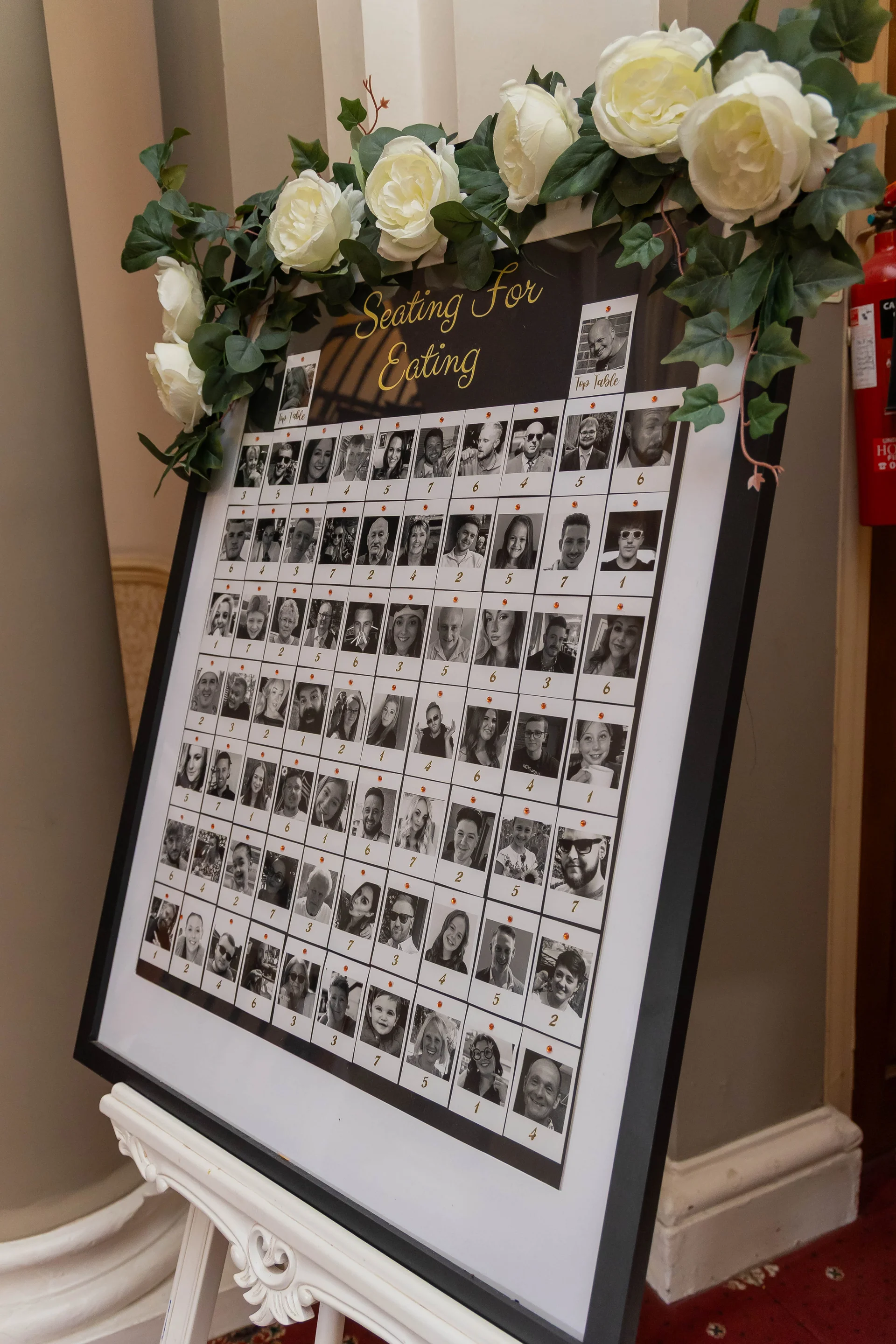 A creative wedding seating plan display featuring rows of black and white guest photographs, decorated with white roses and trailing greenery on an easel.