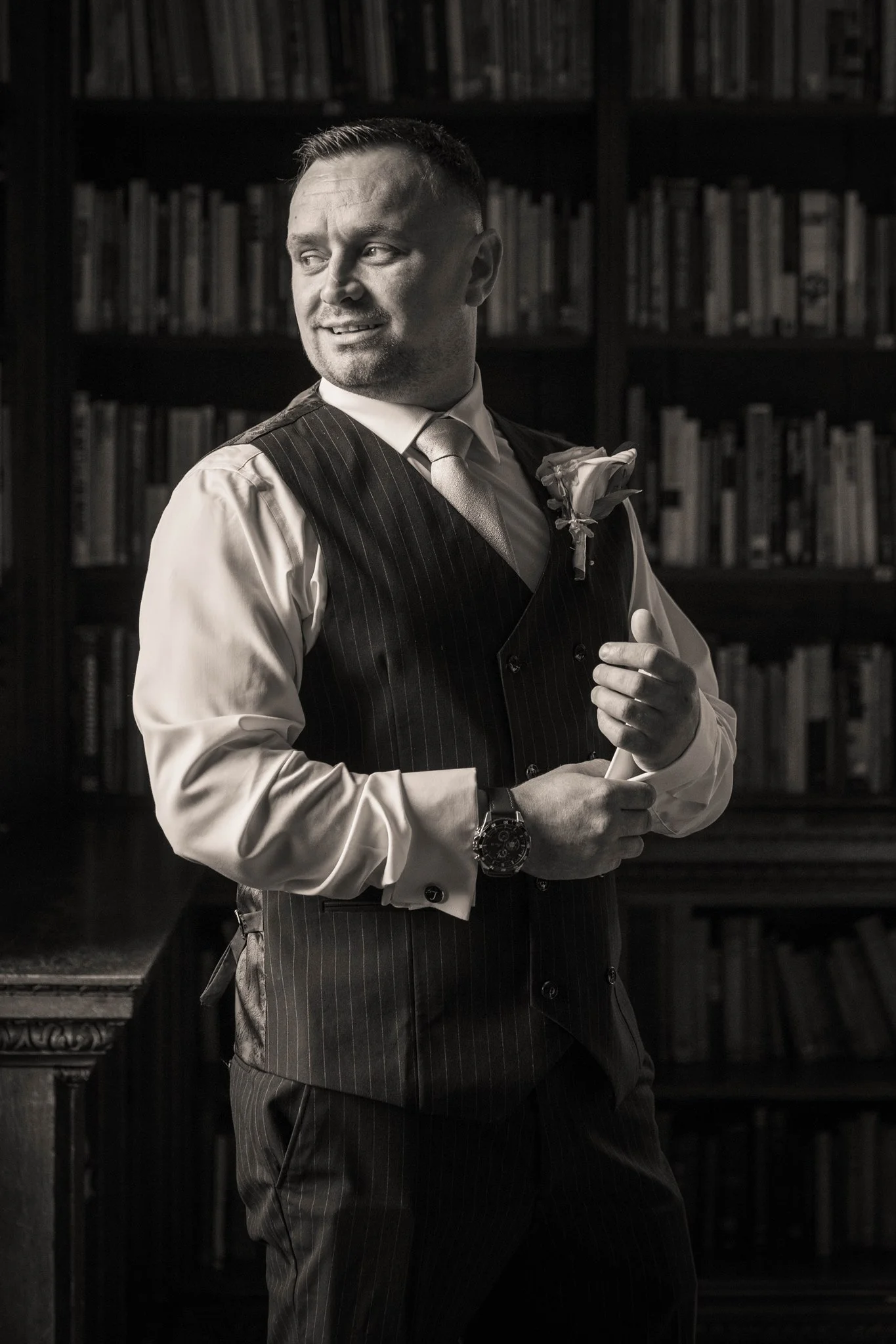A handsome groom in a pinstriped waistcoat adjusts his cufflinks inside a library with floor-to-ceiling bookshelves in this classic black and white portrait.