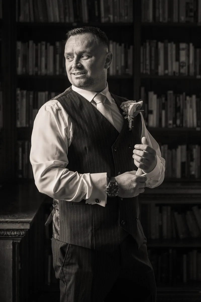 A handsome groom in a pinstriped waistcoat adjusts his cufflinks inside a library with floor-to-ceiling bookshelves in this classic black and white portrait.