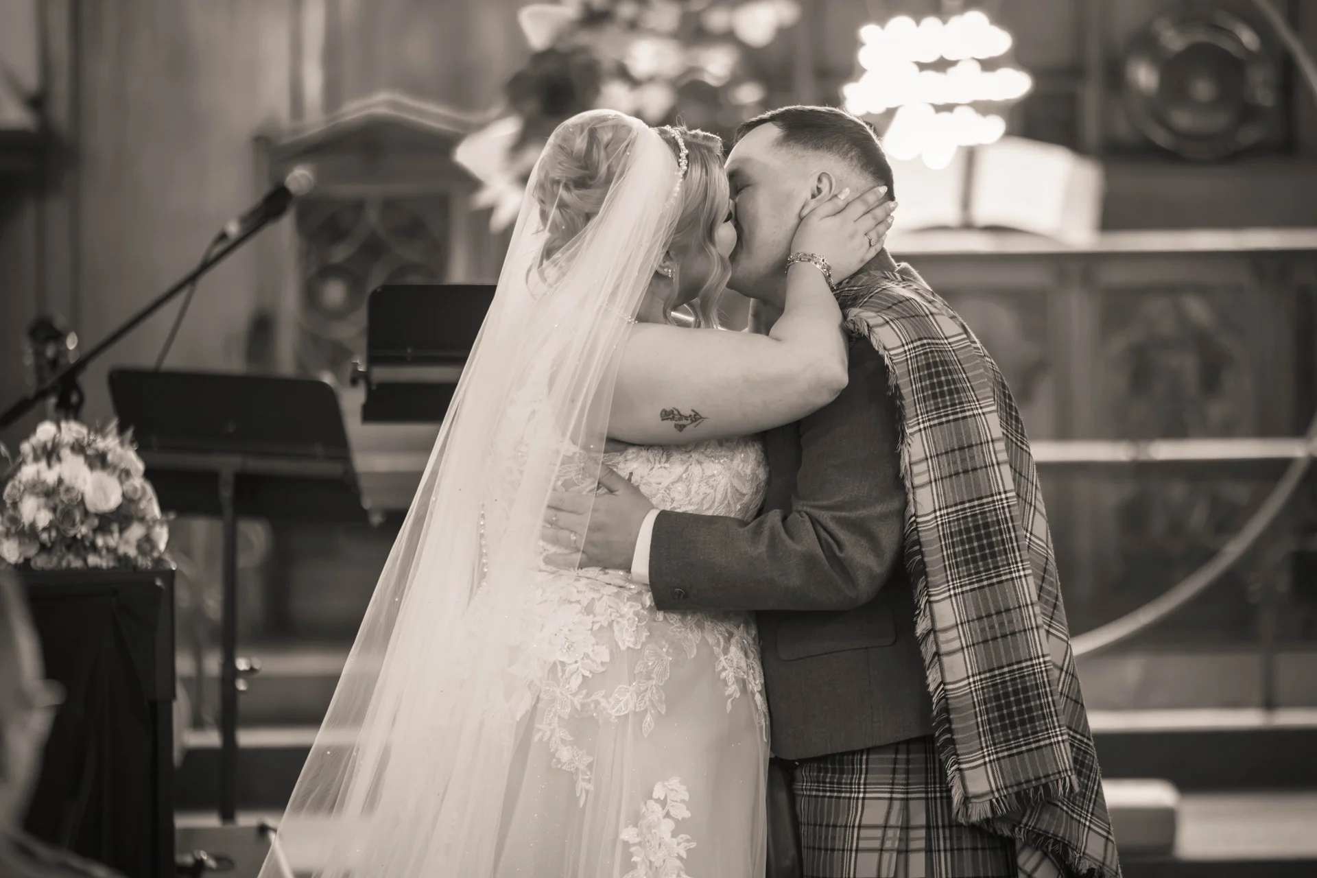 A newly married couple share their first kiss at the altar during a traditional church ceremony, with the groom wearing a tartan kilt and plaid sash.