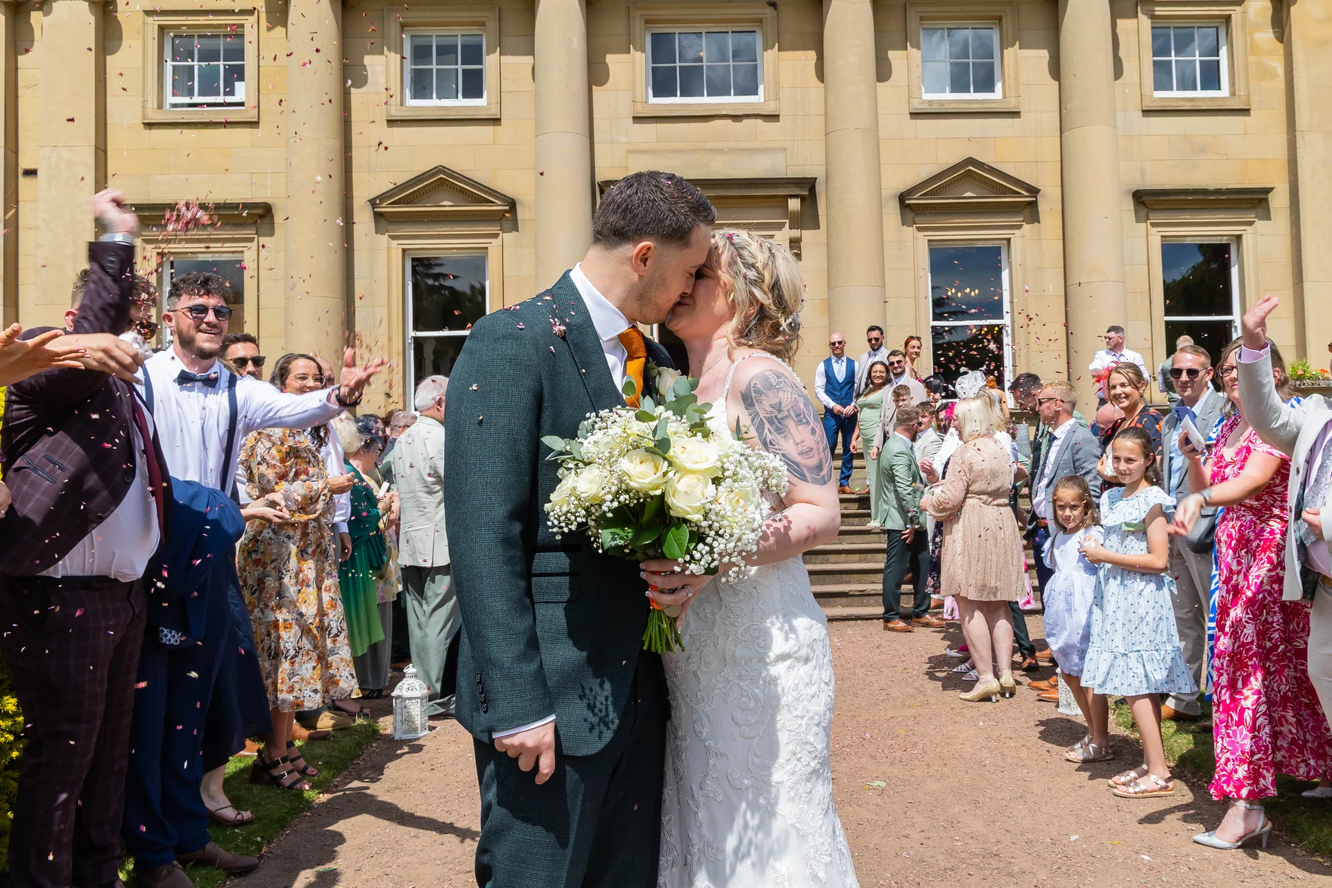 Newlyweds share a romantic kiss outside a grand country house as cheering guests shower them with colorful confetti.