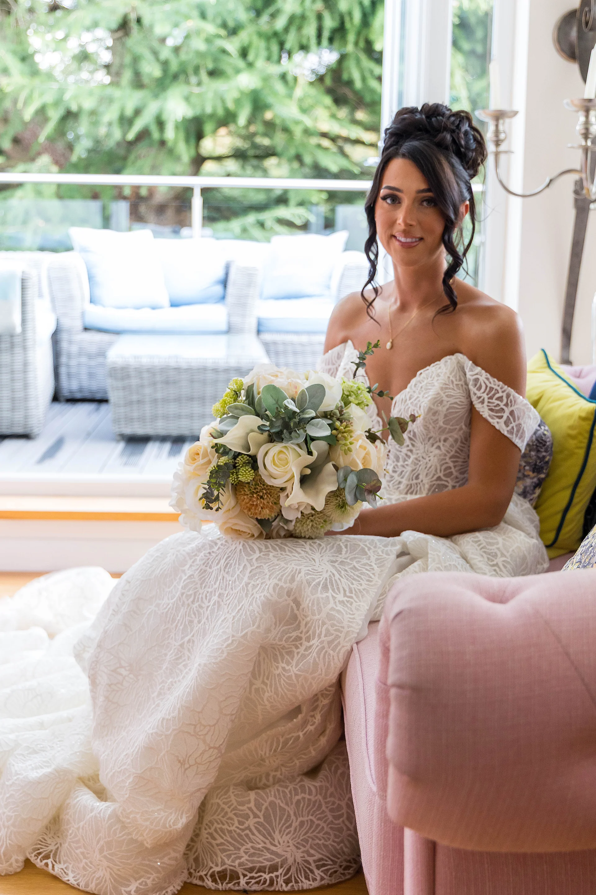 A smiling bride holding a white and green hand-tied bouquet sits gracefully indoors near a large window wearing an off-the-shoulder lace wedding dress and an intricate updo.
