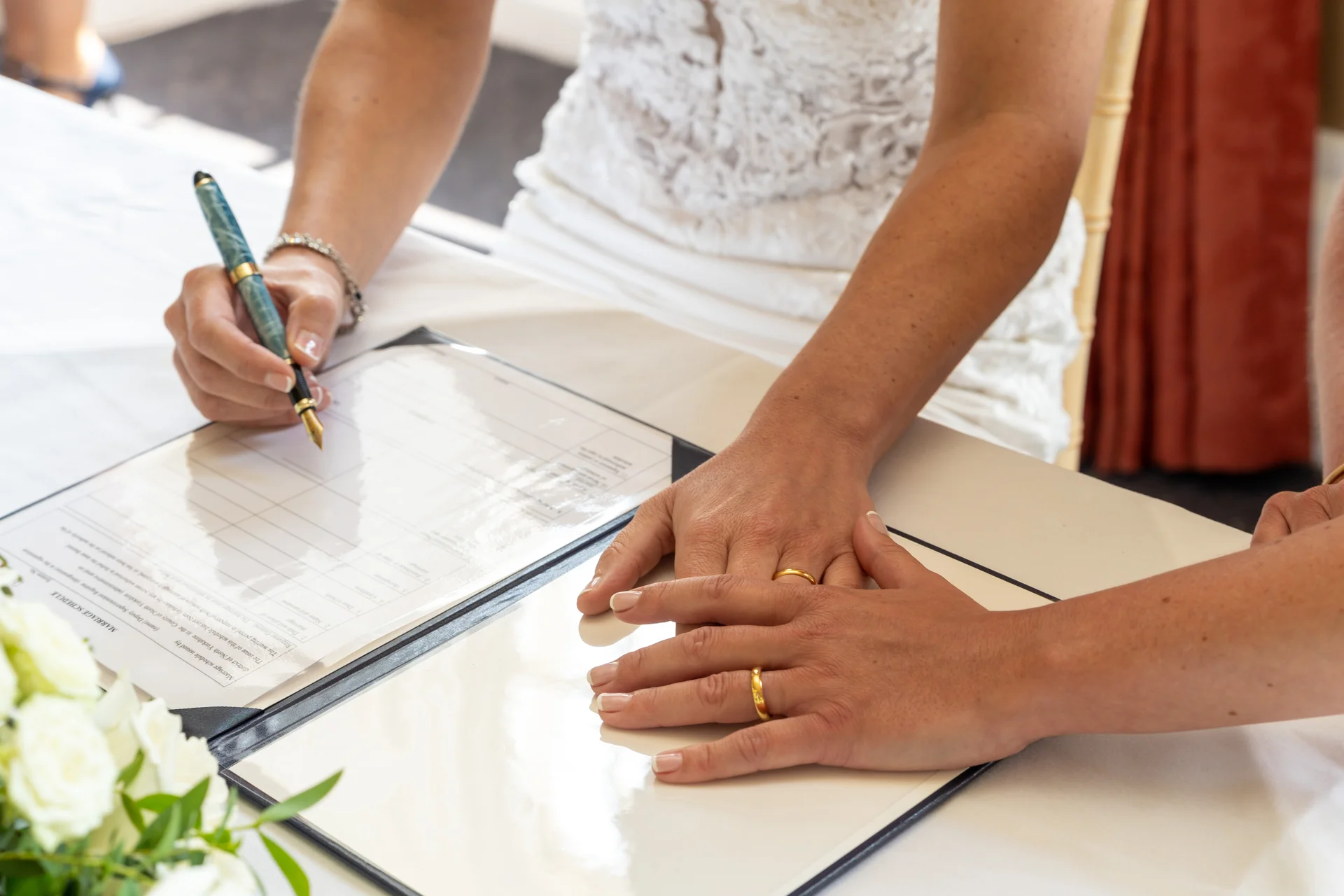 Close-up shot of a bride signing the marriage schedule while two hands rest gently together displaying their wedding rings.