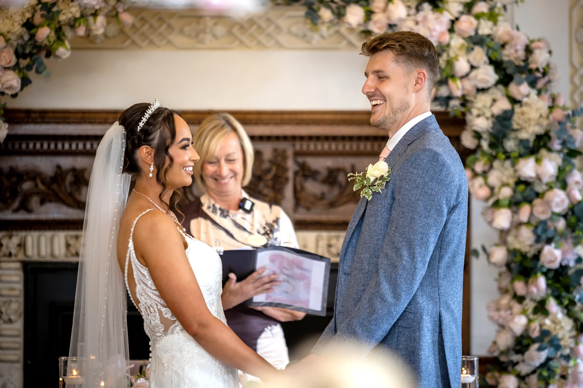 A joyful bride and groom exchange vows during a ceremony framed by a lush floral arch inside a historic venue.