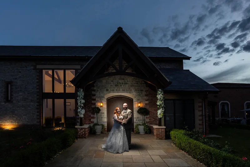 A newlywed couple embracing under the illuminated entrance porch of a striking stone barn venue during twilight.