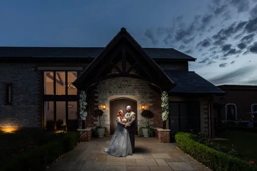 A newlywed couple embracing under the illuminated entrance porch of a striking stone barn venue during twilight.