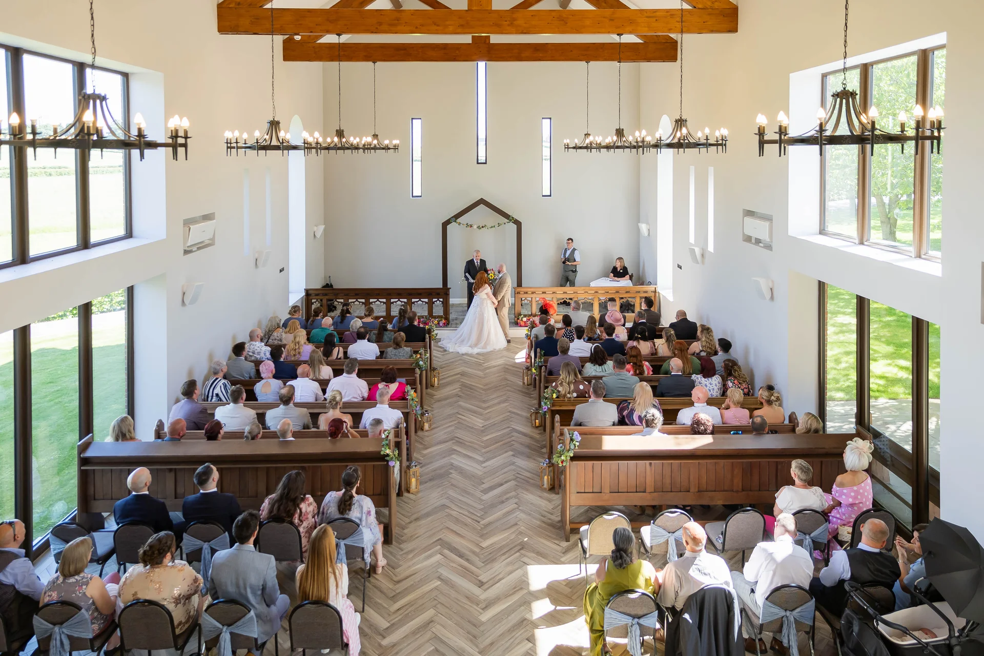 An elevated, wide-angle shot of a bright modern barn wedding ceremony, showing the bride and groom standing before an altar while seated guests fill wooden pews beneath high timber rafters.