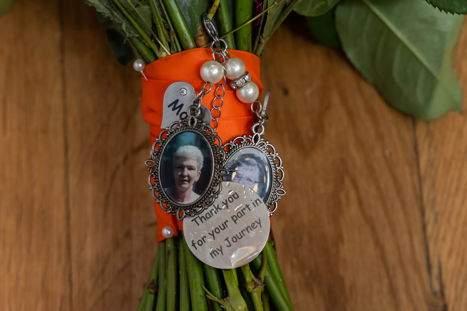 A close up of a hand-tied bridal bouquet wrapped in vibrant orange ribbon featuring memorial photo charms and engraved tokens attached as a touching tribute.