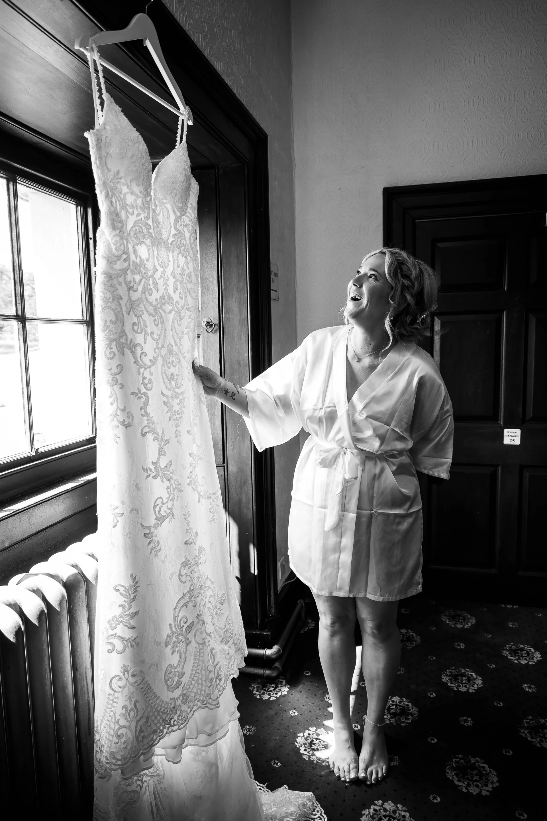 A joyful bride in a white robe stands barefoot, gazing up with excitement at her intricately detailed wedding dress hanging from a dark wooden doorway.