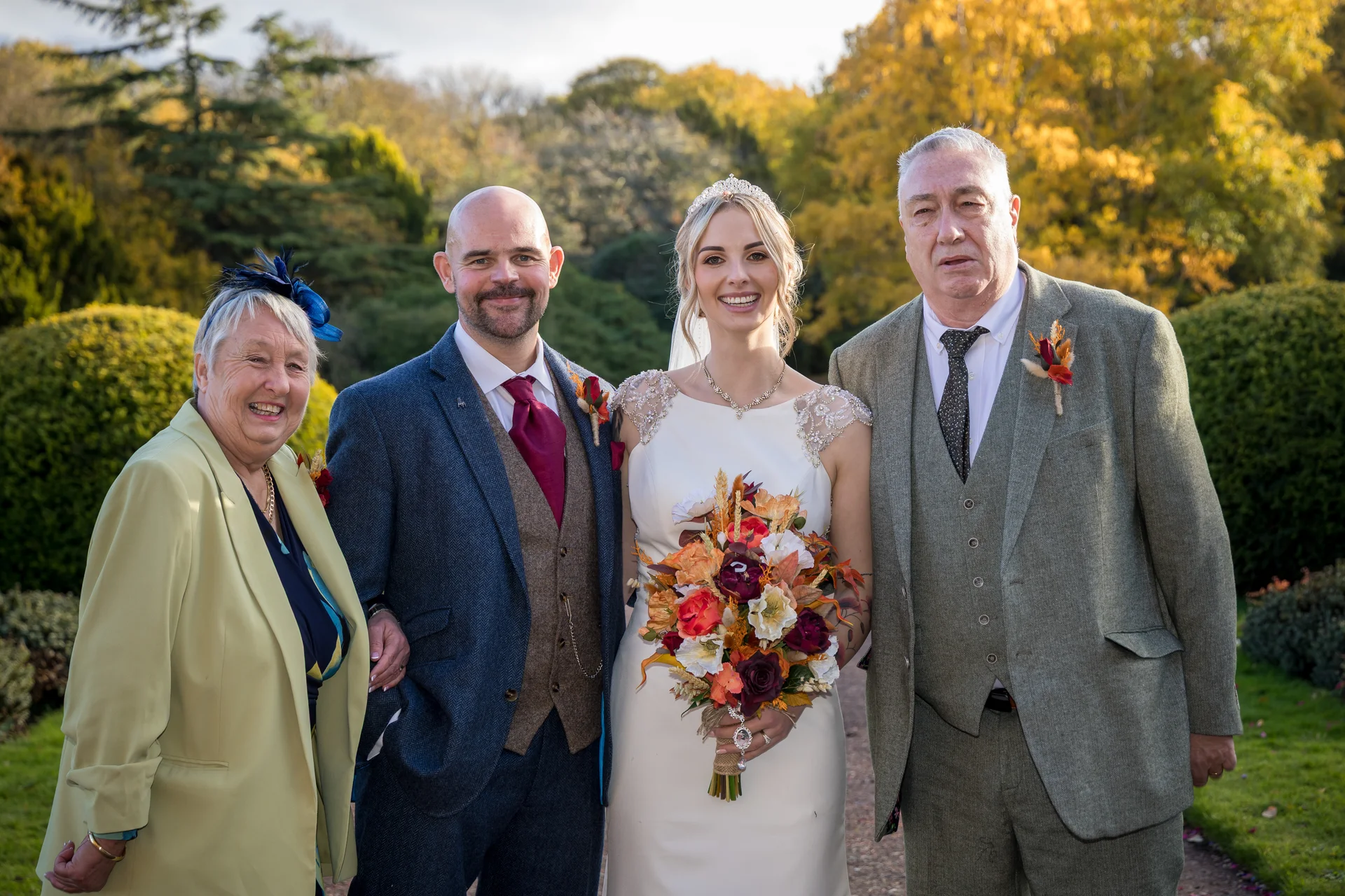 A smiling bride and groom stand outdoors in an autumn garden alongside their family members, surrounded by vibrant fall foliage and warm seasonal tones.
