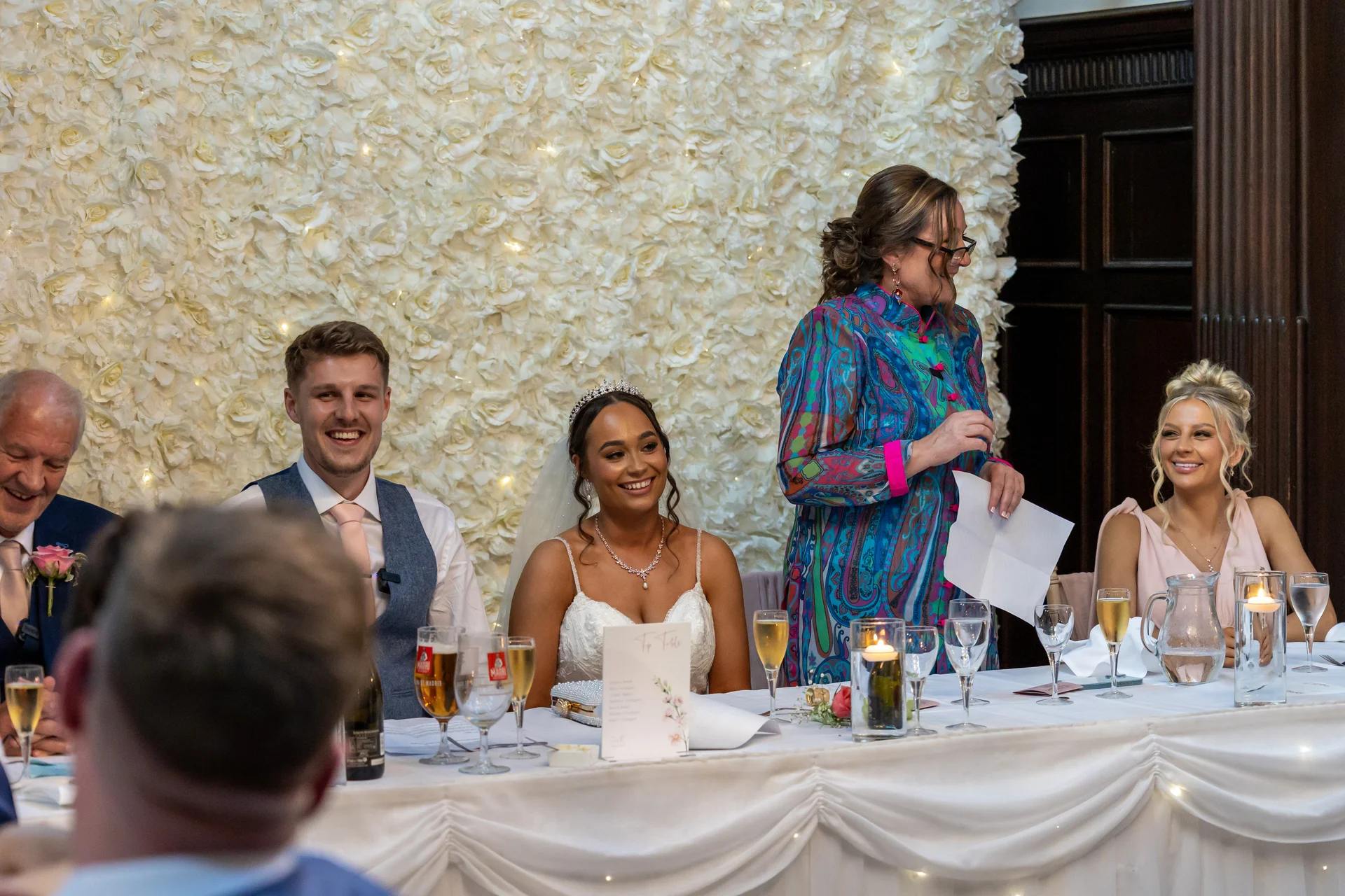 The bride and groom smile seated at the top table during wedding speeches in front of a large white rose wall.