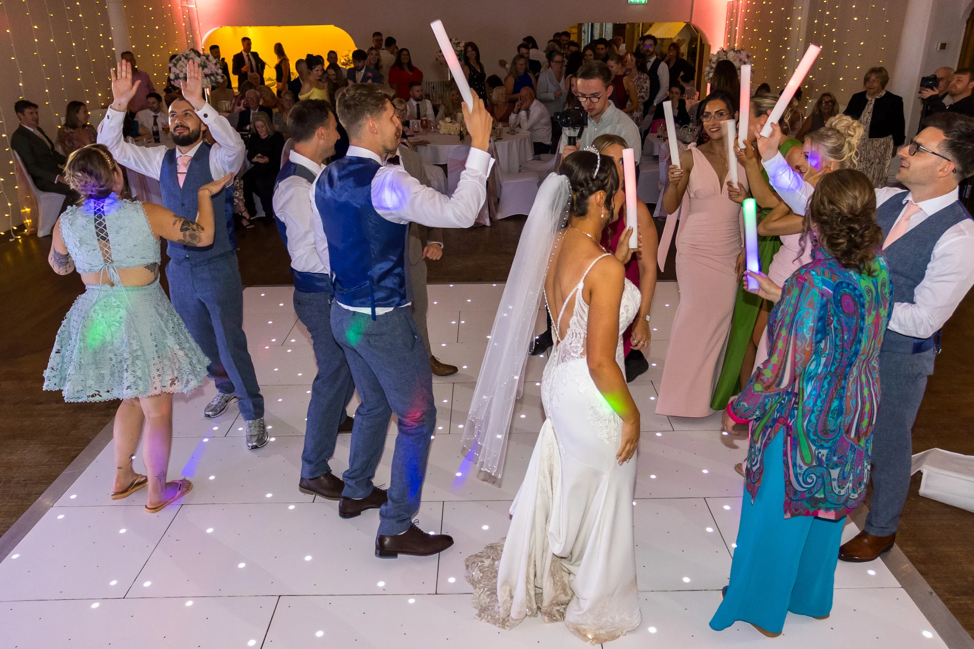 The bride and groom dance with enthusiastic guests on an illuminated white dance floor while holding glowing foam batons during the evening reception.