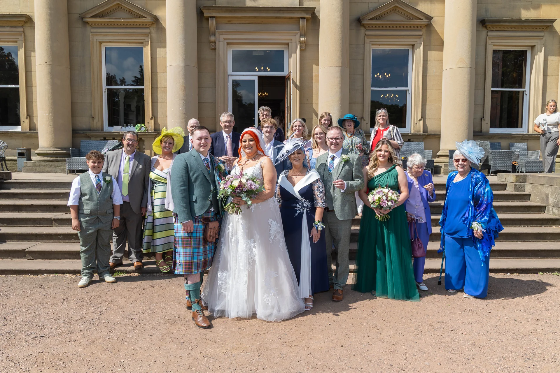 A large wedding party and family members pose for a formal group photograph on the grand stone steps outside a stately home.