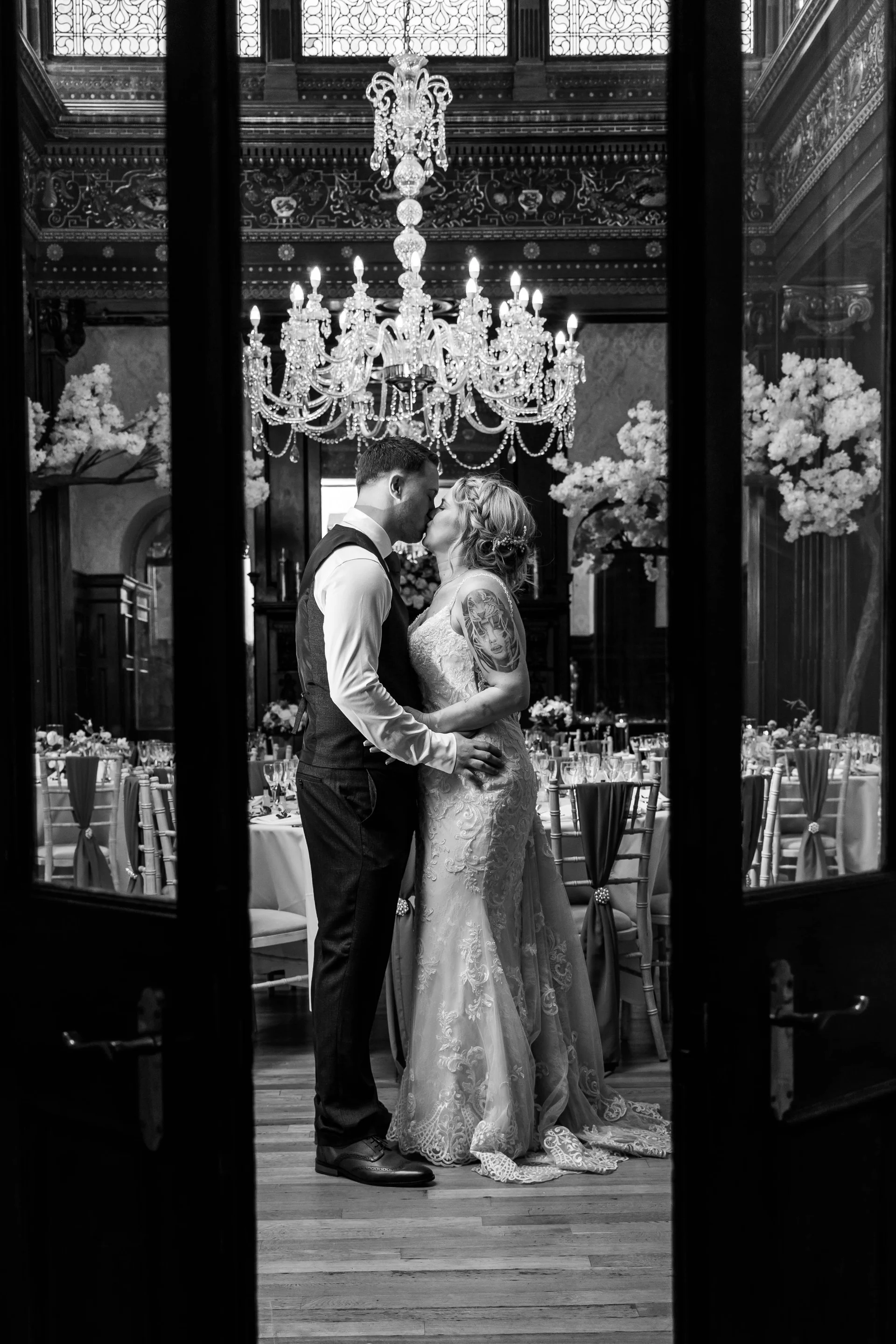 A newlywed couple share a tender kiss inside an opulent ballroom illuminated by a grand crystal chandelier, viewed through open double doors.
