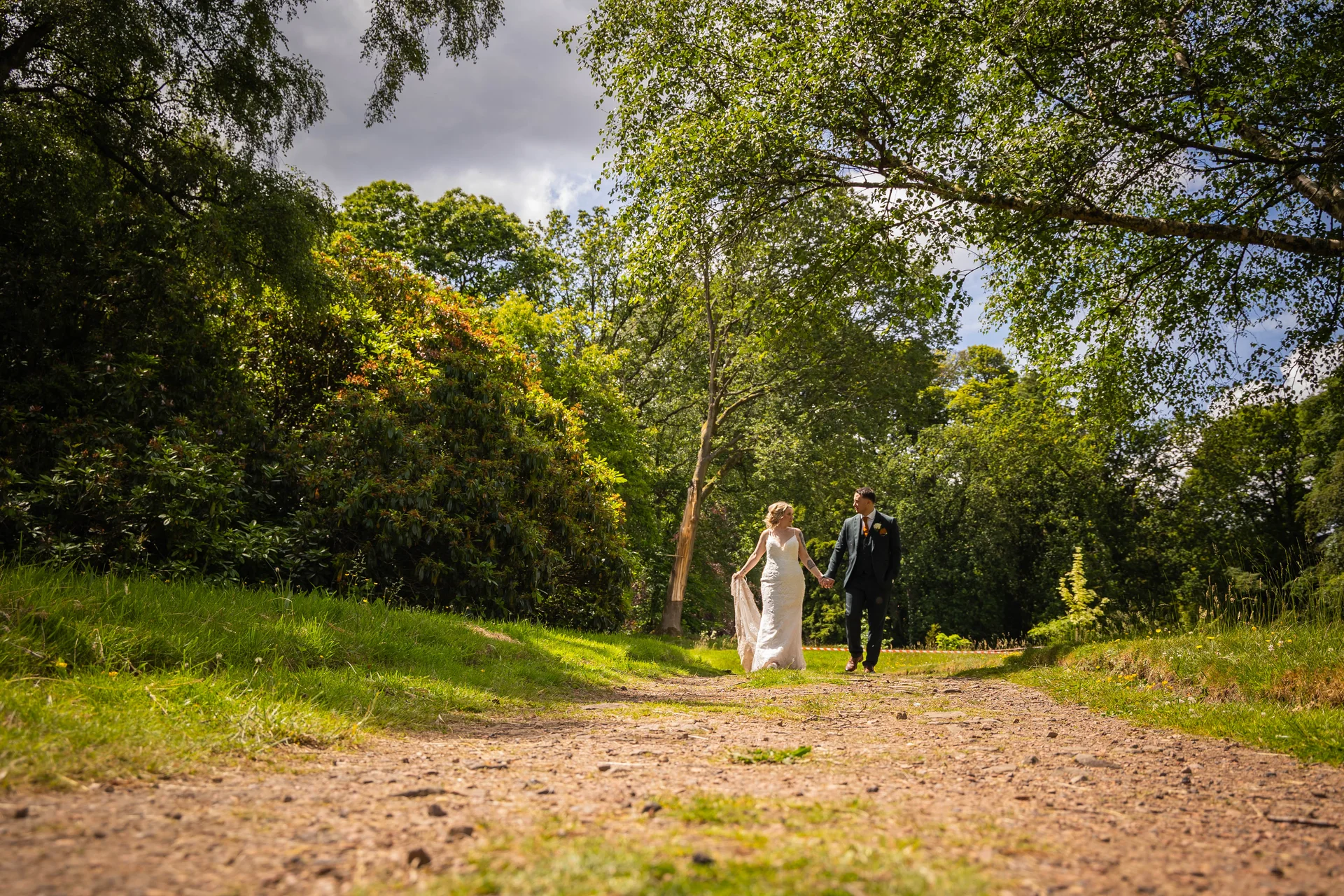 A newlywed couple holds hands while walking along a sunlit dirt path surrounded by lush green woodland and trees.